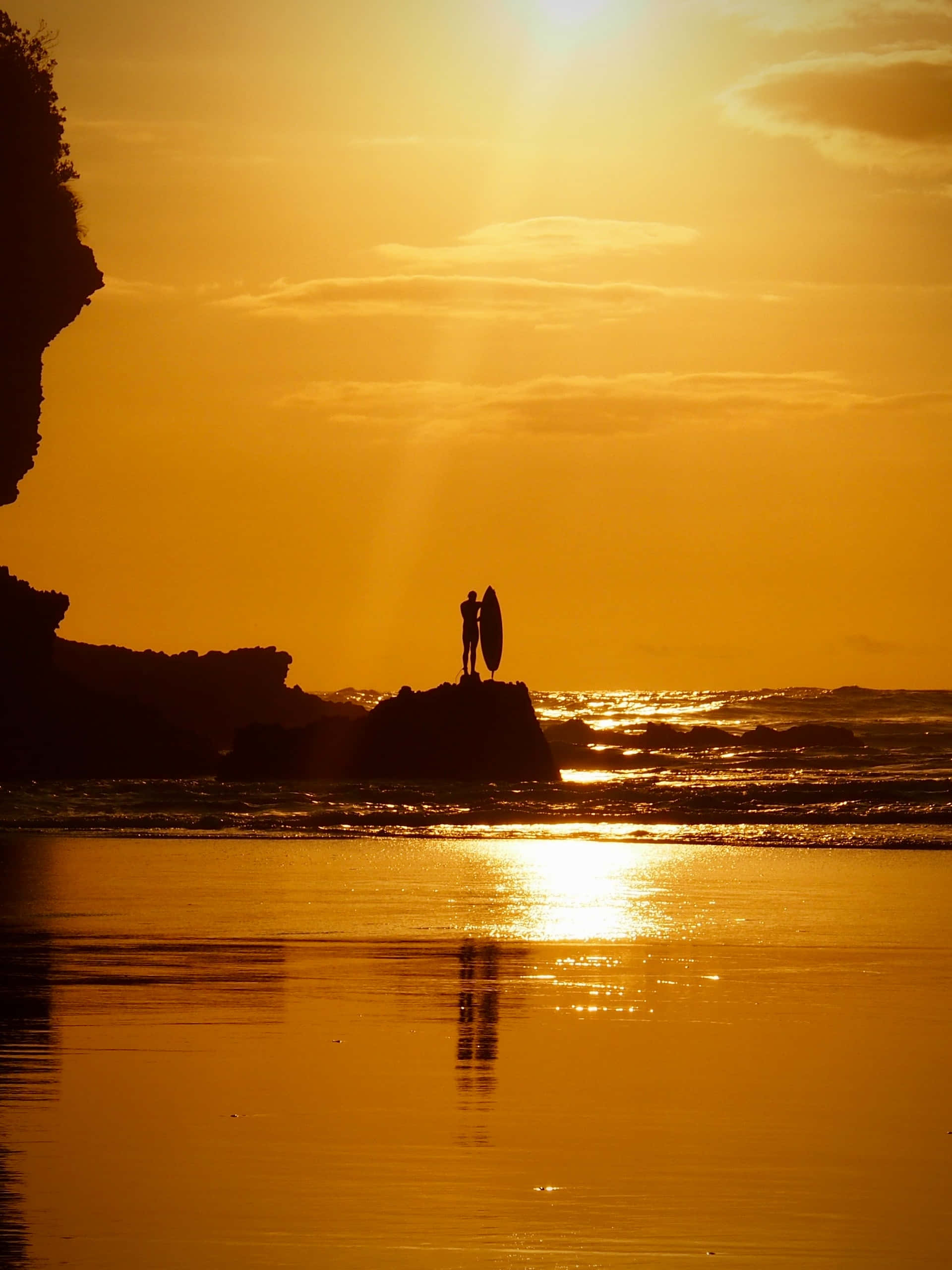 Yellow Beach 桌布 壁紙 — 金色夕陽下，衝浪者剪影站在岩石上，倒影映在濕潤沙灘和海浪上。