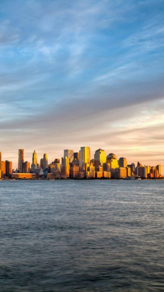 日落 桌布 壁紙 — 金色夕陽以溫暖的光芒沐浴紐約市天際線，並倒映在下方平靜的水面上。