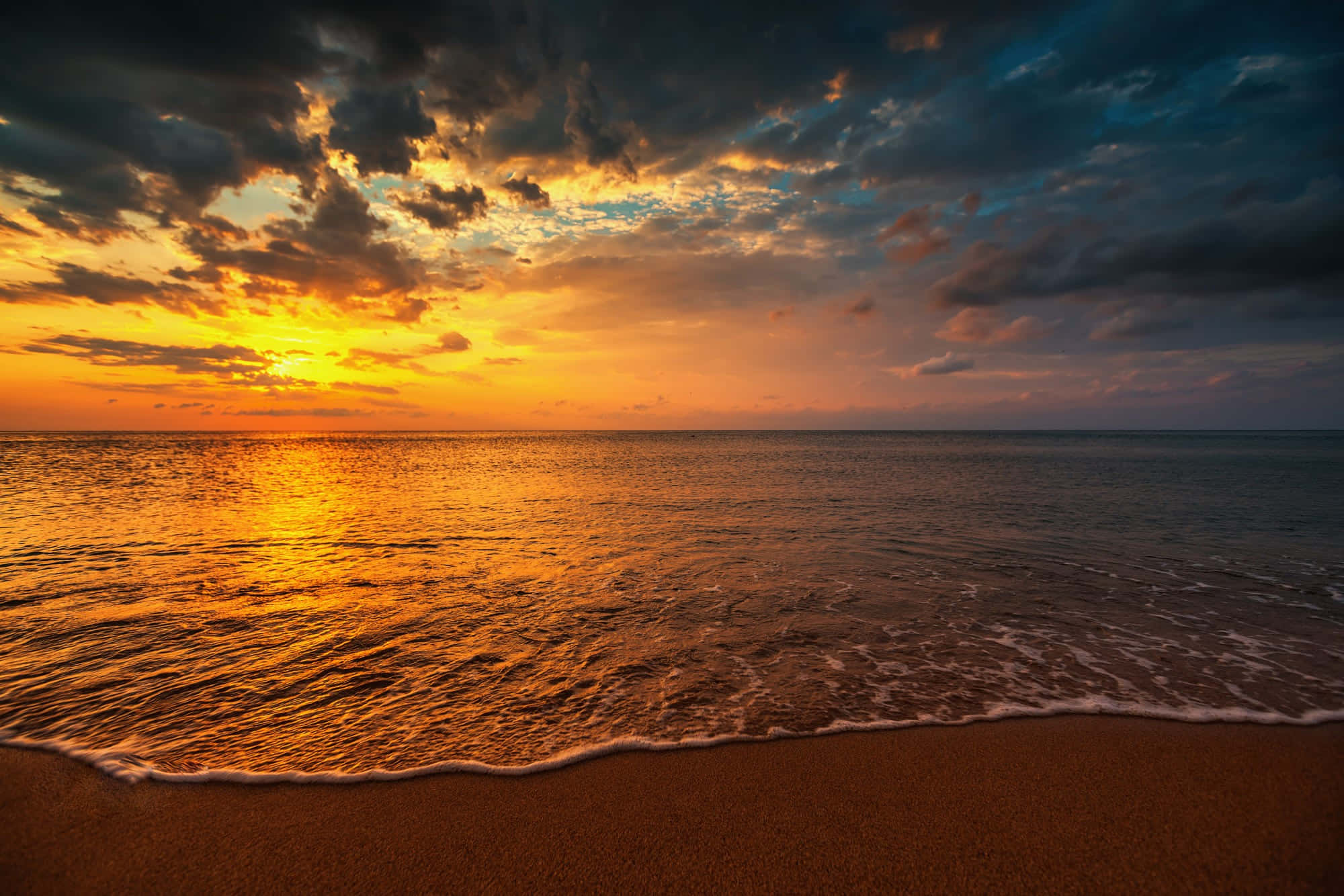 Sunset Ocean 桌布 壁紙 — 金色夕陽以火熱色調描繪海洋和天空，海浪在戲劇性的雲層下輕輕親吻沙灘。