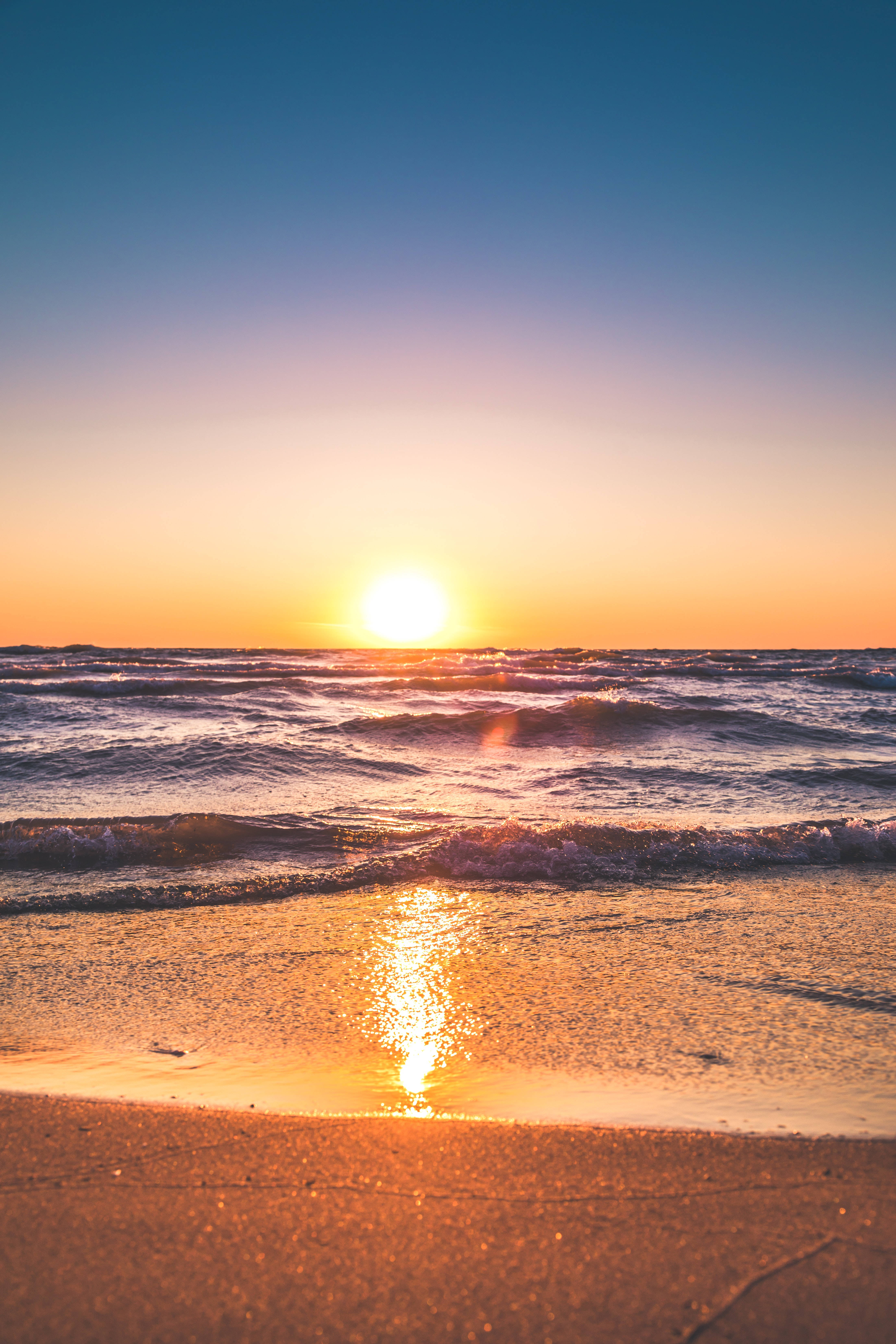 日落 桌布 壁紙 — 金色夕陽在海浪上閃耀，沖刷著沙灘，天空呈現鮮豔的橙色和藍色漸變。