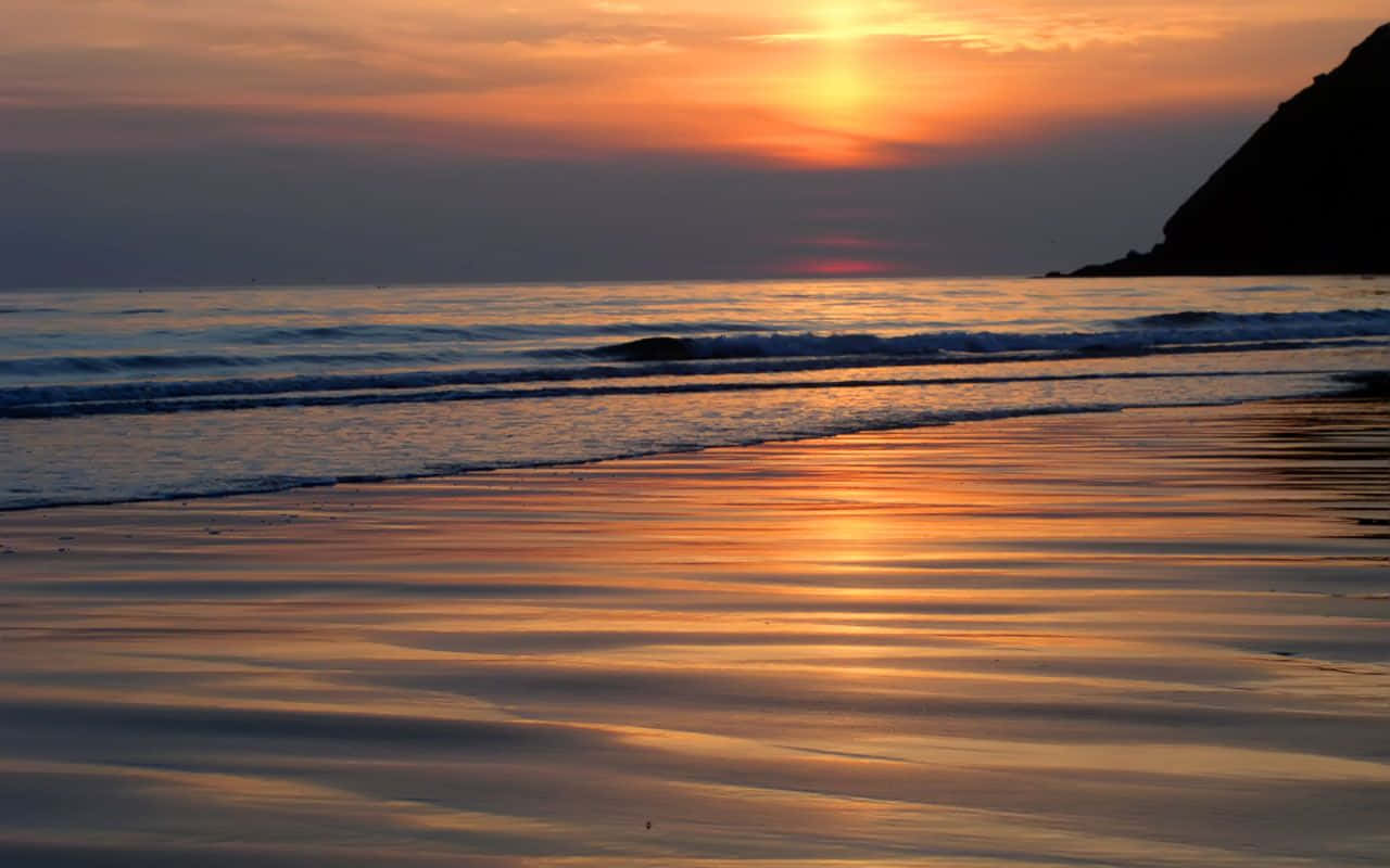 Sunset View 桌布 壁紙 — 金色夕陽在濕沙上閃耀，溫柔的海浪在黑暗懸崖剪影旁翻滾。