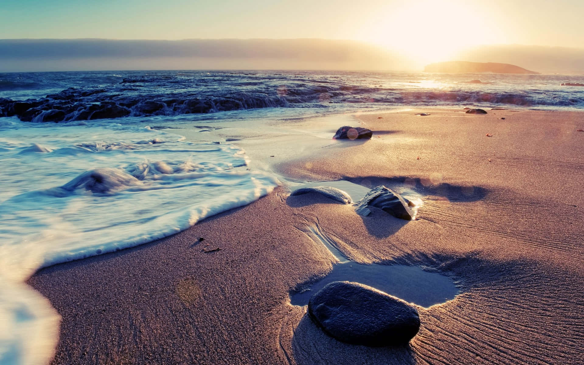 Beach Waves 桌布 壁紙 — 金色夕陽將溫暖光線灑在濕沙上，溫柔的海浪拍打著散落石塊的岩石海灘。