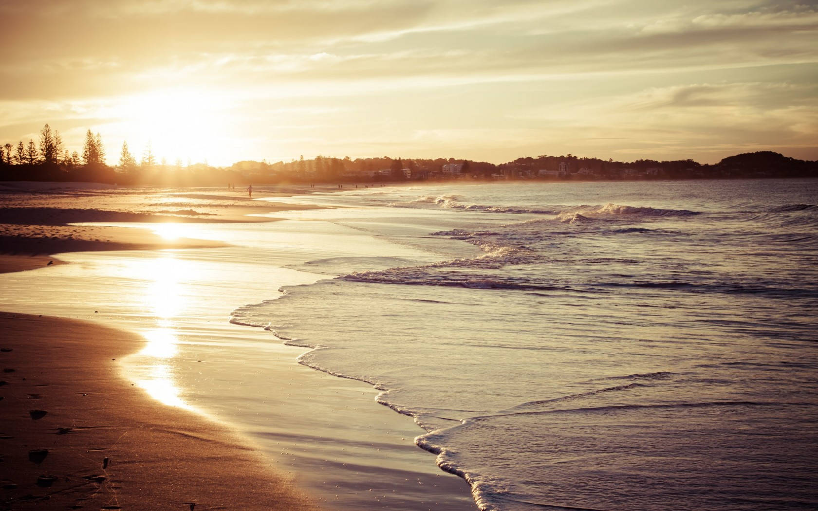 Beach Desktop 桌布 壁紙 — 金色夕陽將溫暖的光線投射在濕沙上，溫柔的海浪拍打著寧靜的海灘，遠處有樹木和一個孤獨行走的人影。