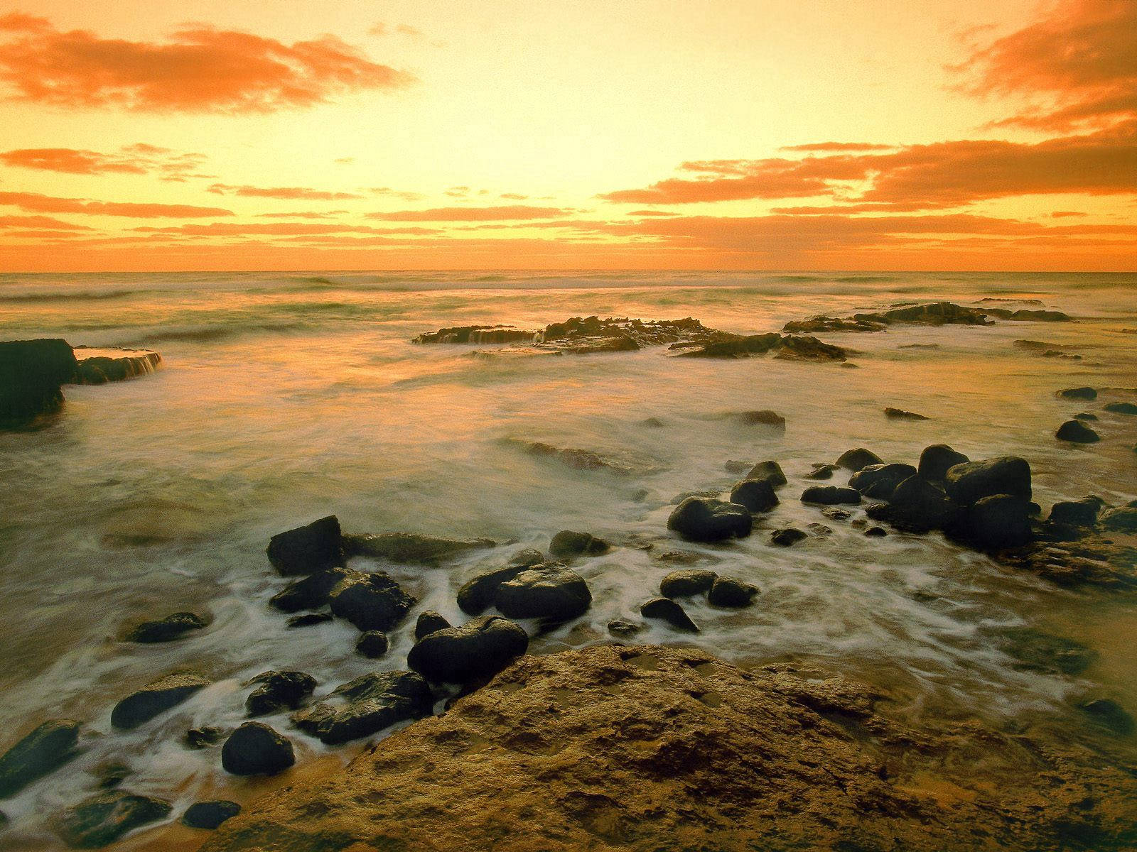  壁紙 — 金色夕陽染紅海洋，海浪輕輕拍打黑色岩石，營造出寧靜的海岸景色。