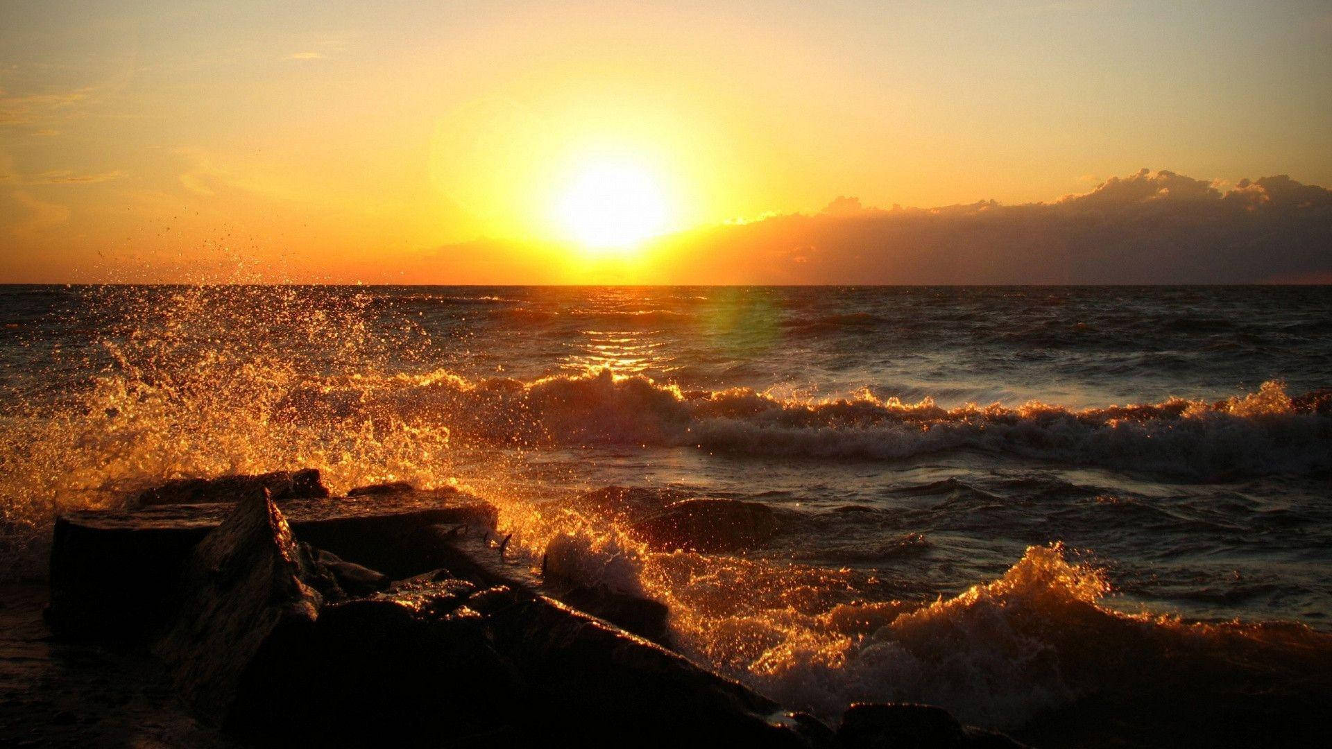 日落 桌布 壁紙 — 金色夕陽沐浴在洶湧的海浪拍打黑色岩石上，散發出火熱的橙色和黃色光芒。