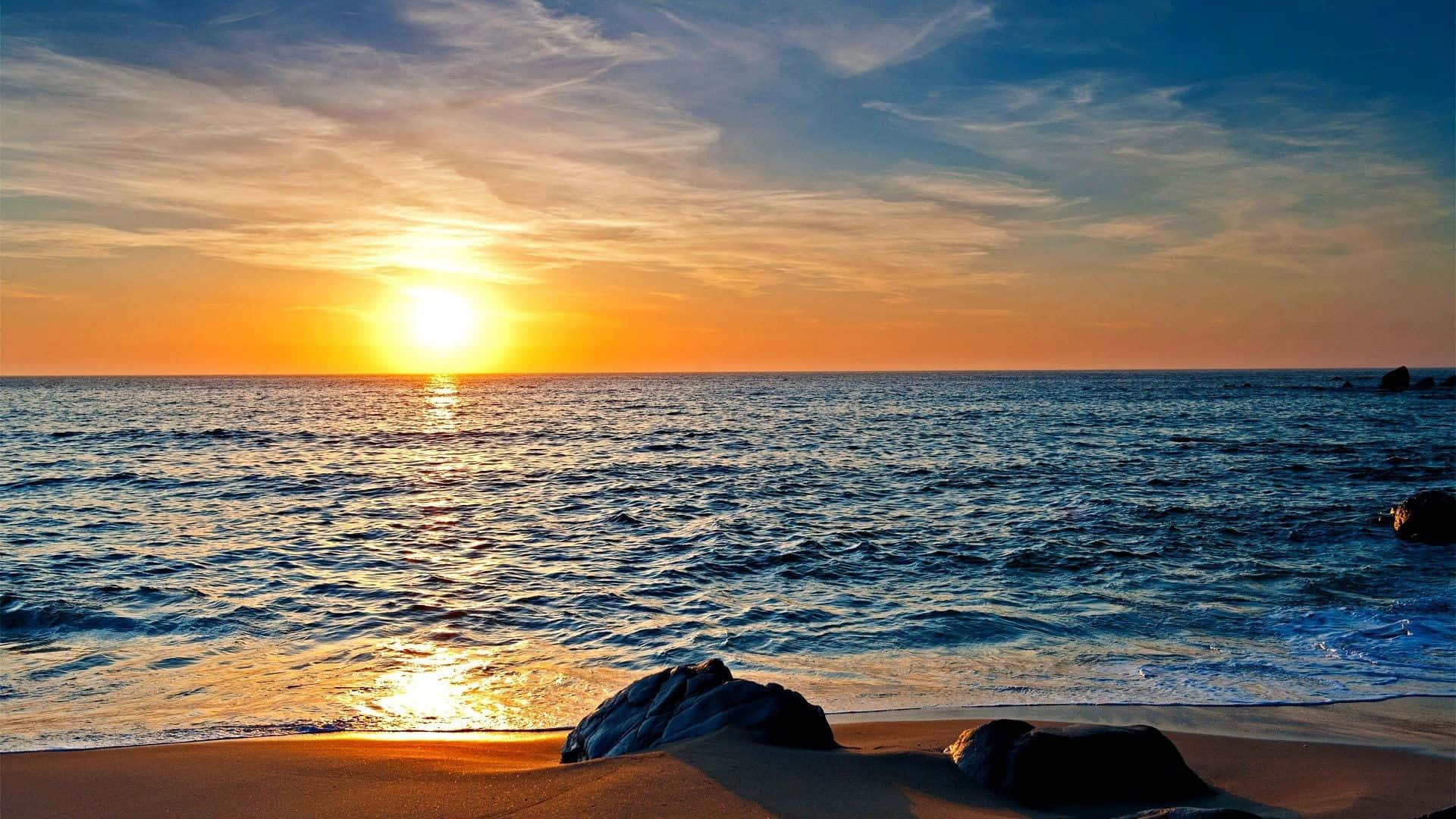 Sunset View 桌布 壁紙 — 金色夕陽照耀在平靜的海浪上，海浪沖刷著沙灘，前景有深色岩石。
