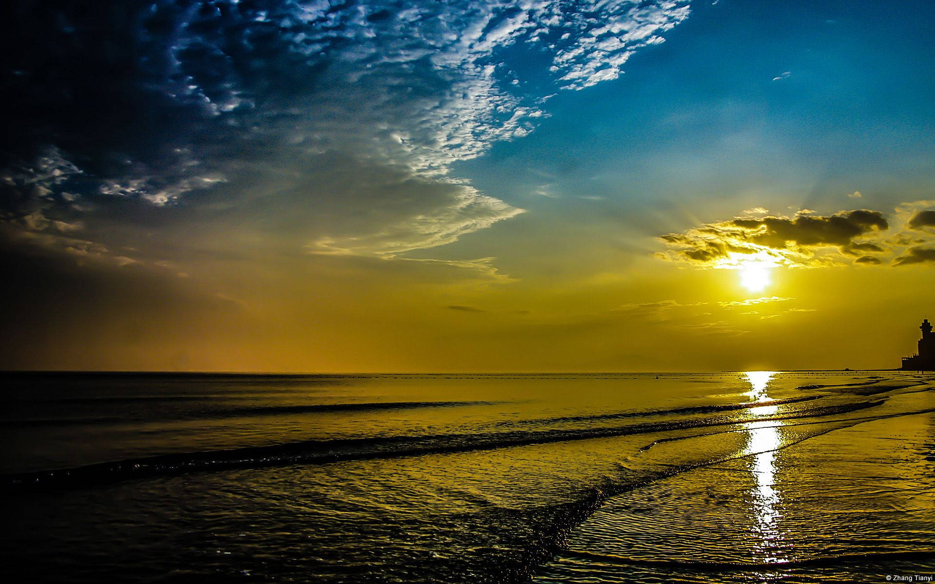 日出 桌布 壁紙 — 金色夕陽照耀在海浪上，伴隨著戲劇性的雲彩和遠處燈塔的剪影。