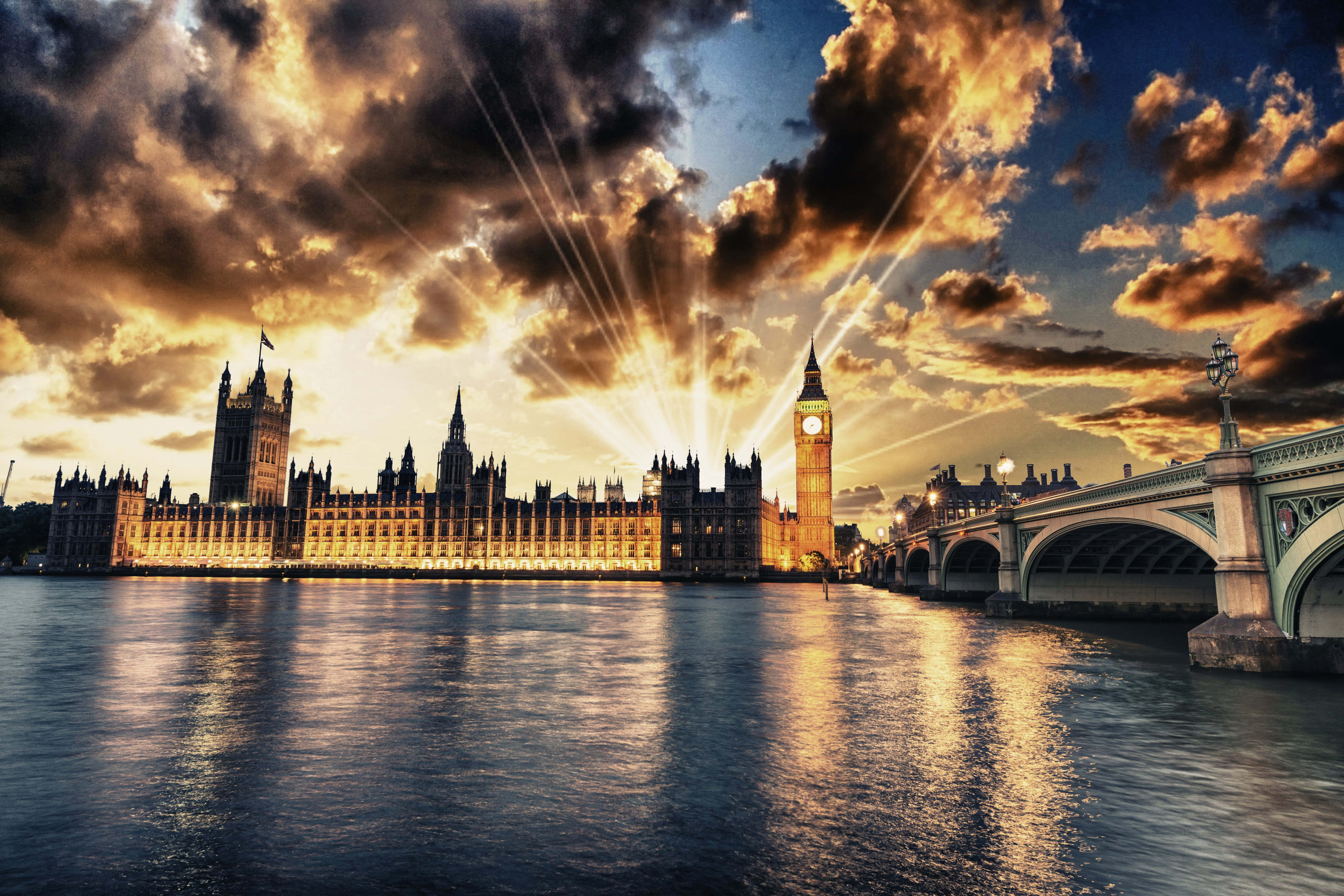 UK 桌布 壁紙 — 金色夕陽穿過戲劇性的雲層，照耀在倫敦標誌性的大笨鐘和國會大廈上，倒映在泰晤士河中。