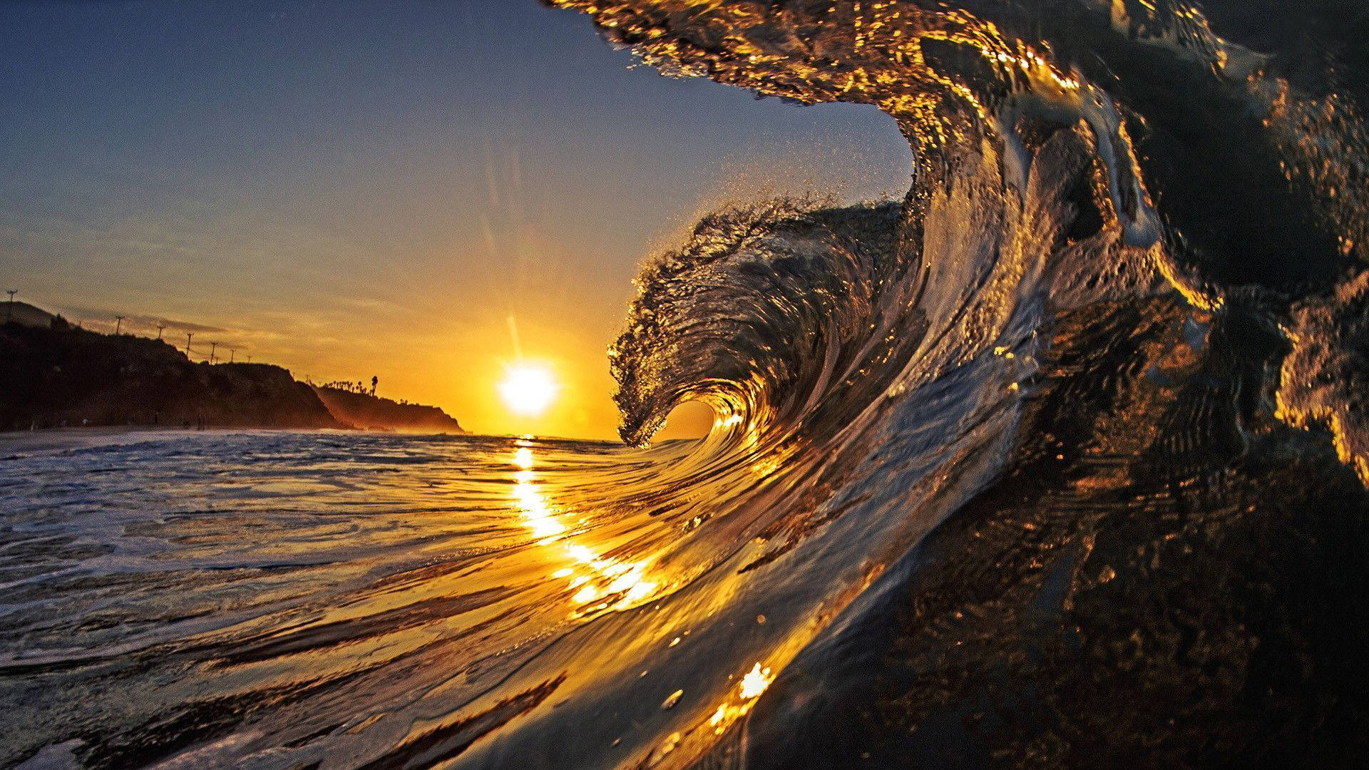 海灘日落 桌布 壁紙 — 金色夕陽穿過破碎海浪的筒身閃耀，背景有閃爍海水和暗色懸崖。