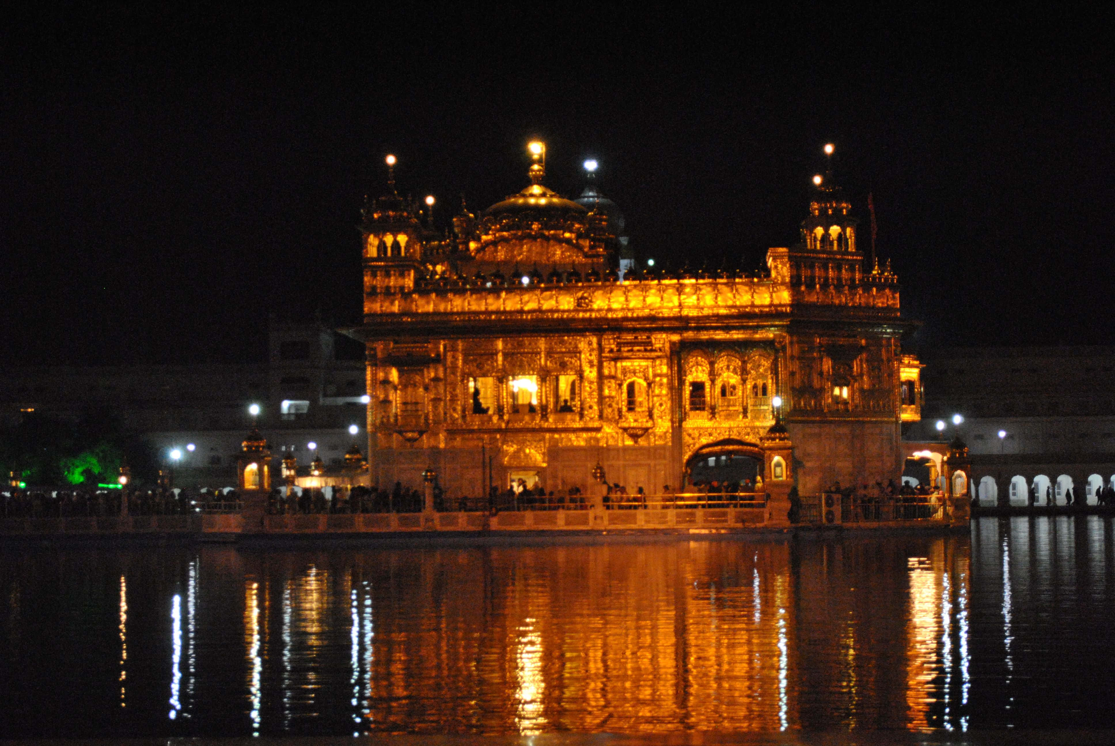 Golden Temple 桌布 壁紙 — 金色寺廟在夜晚被溫暖的燈光照亮，美麗地倒映在平靜的周圍水面上。