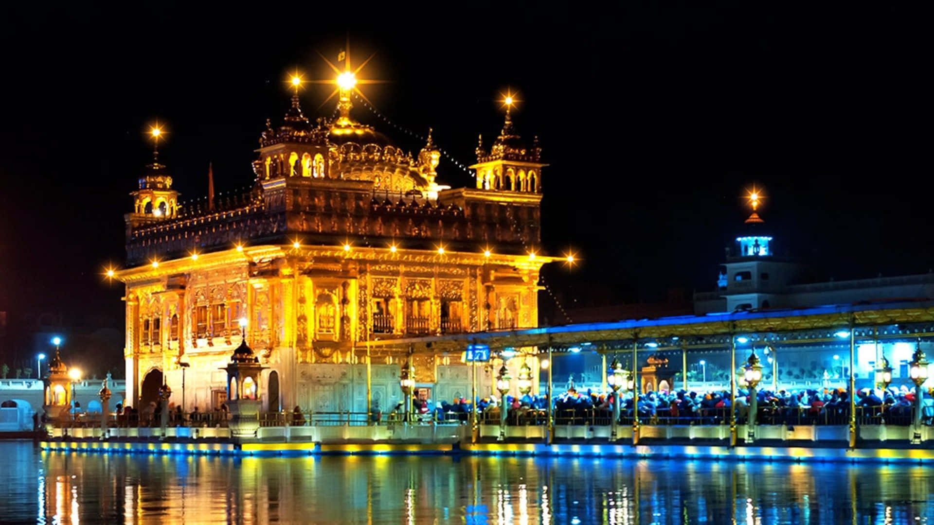 Golden Temple 桌布 壁紙 — 金色寺廟在夜晚被照亮，鮮豔燈光倒映在平靜的水面上，周圍擠滿人群。