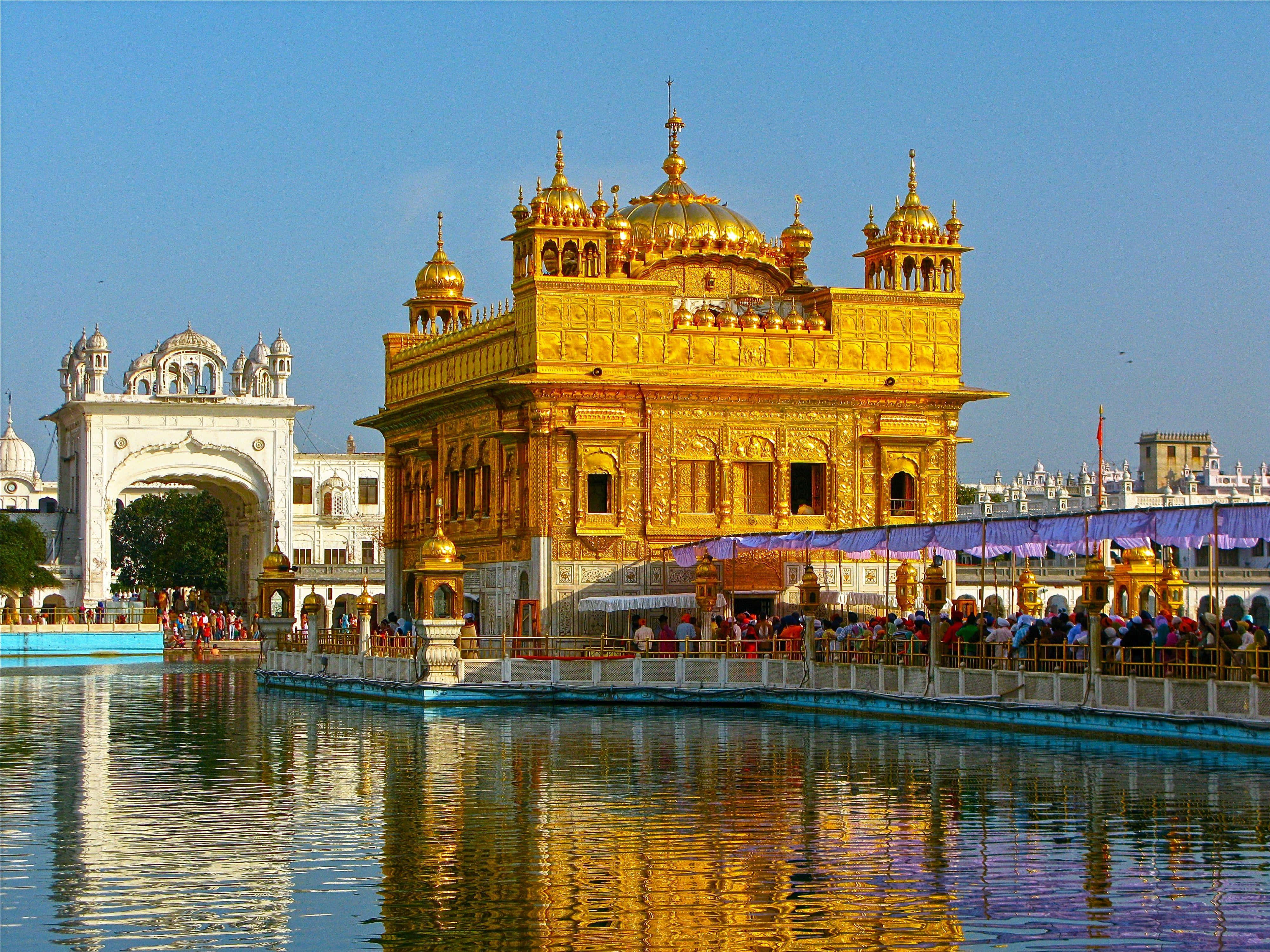 Golden Temple 桌布 壁紙 — 金色寺廟在明亮陽光下閃耀，倒映在平靜的水面上，遊客聚集在其神聖的池塘周圍。