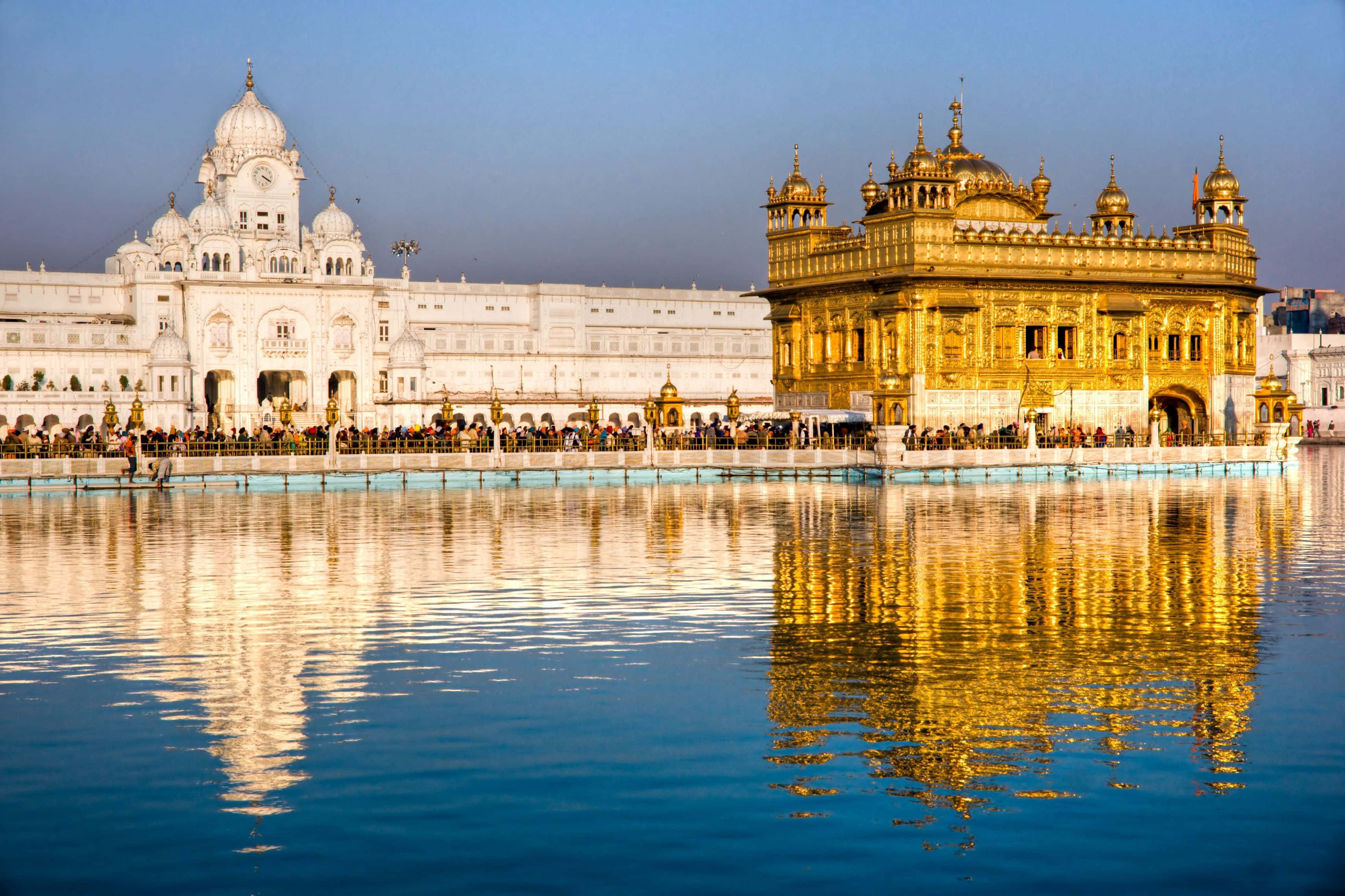 Golden Temple 桌布 壁紙 — 金色寺廟在晴朗天空下閃耀，在平靜的藍色水面上完美倒映，周圍有遊客。