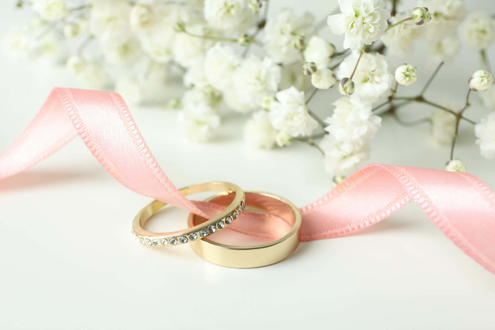 金色戒指與粉紅絲帶婚禮背景 壁紙 — 兩枚金色結婚戒指纏繞著柔軟的粉紅色絲帶，背景是精緻的白色滿天星花朵。