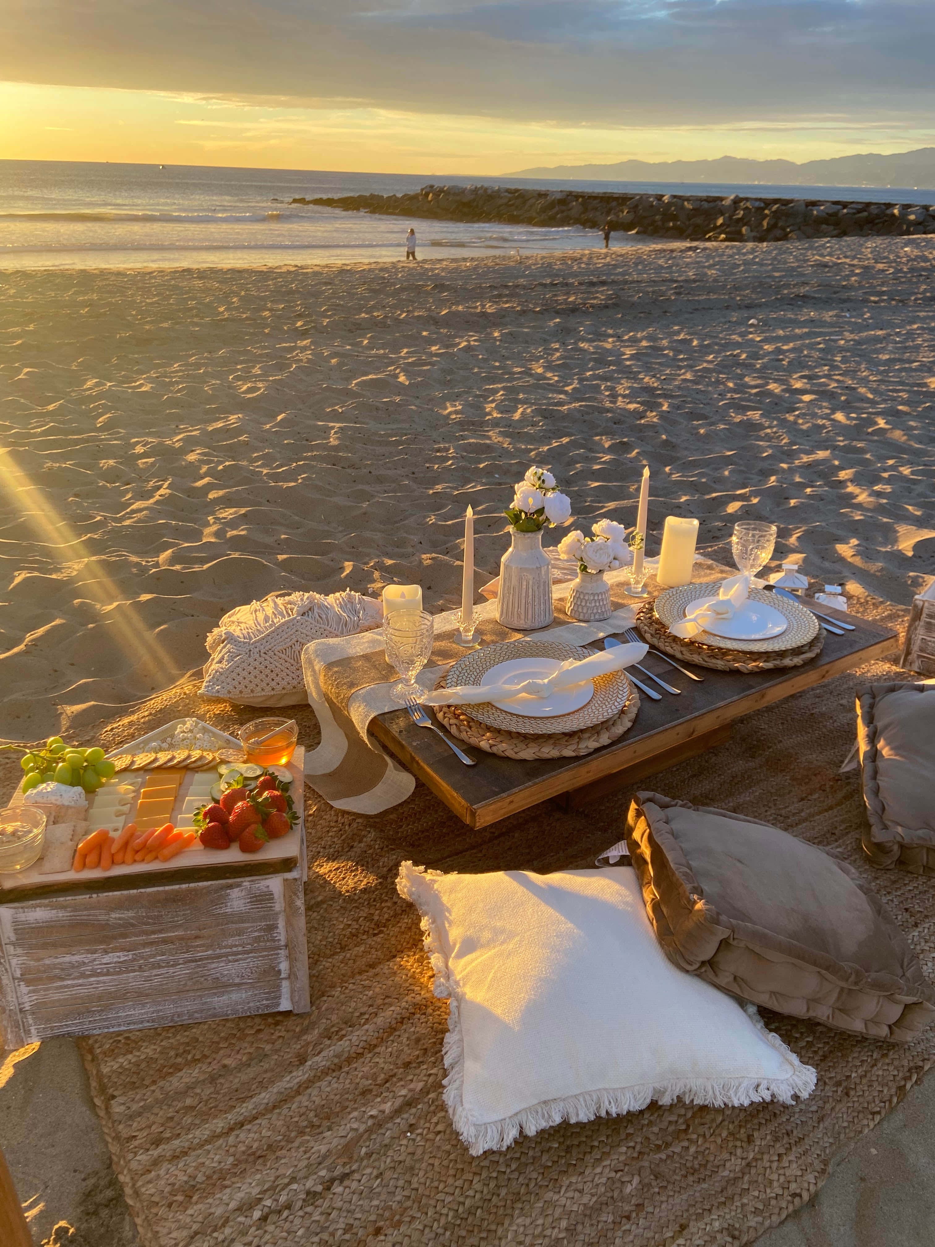 Beach Picnic 桌布 壁紙 — 金色日落下的浪漫沙灘野餐，設有蠟燭、碟子和新鮮水果在編織墊上。