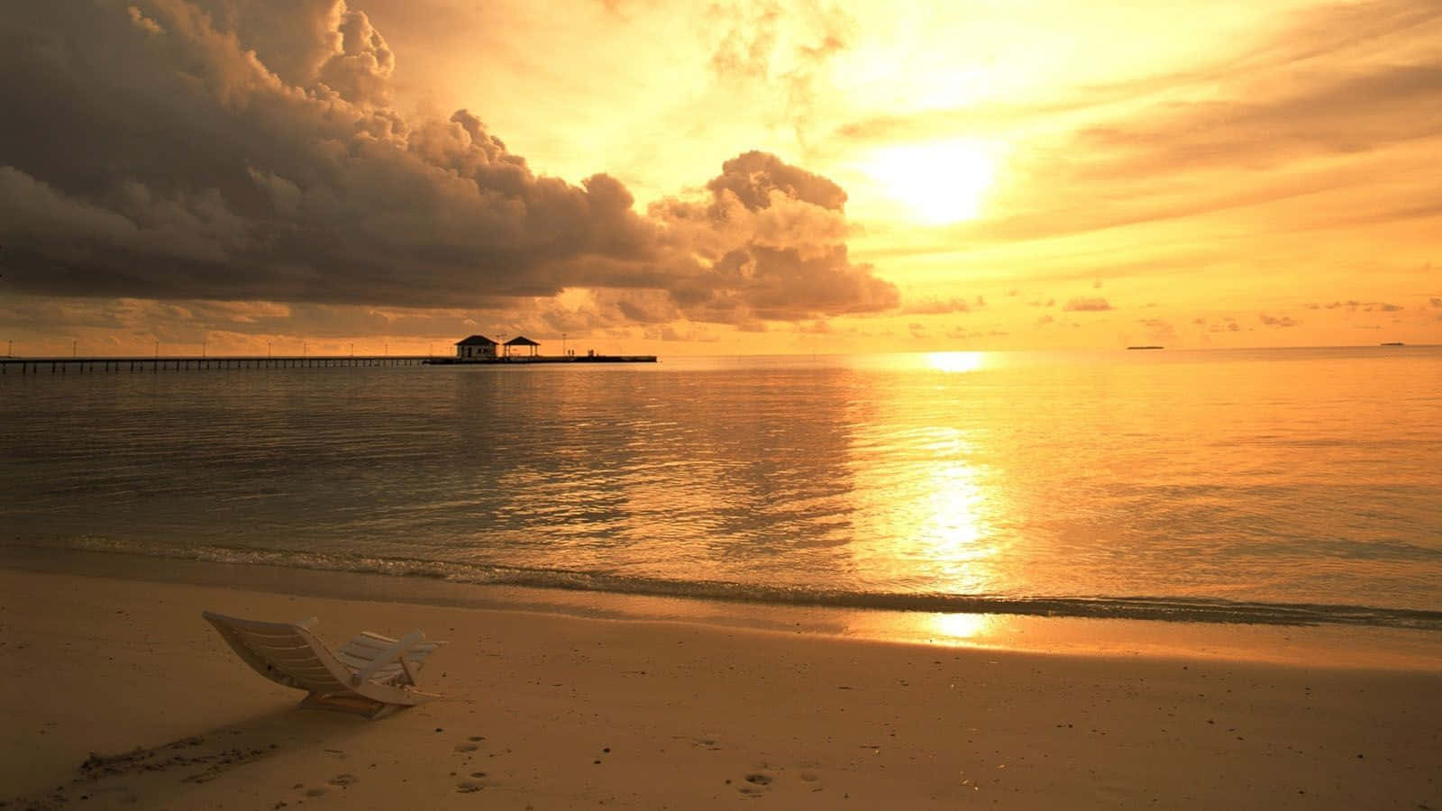 Yellow Beach 桌布 壁紙 — 金色日落映照平靜海洋，孤獨沙灘椅和碼頭在戲劇性雲層中形成剪影。