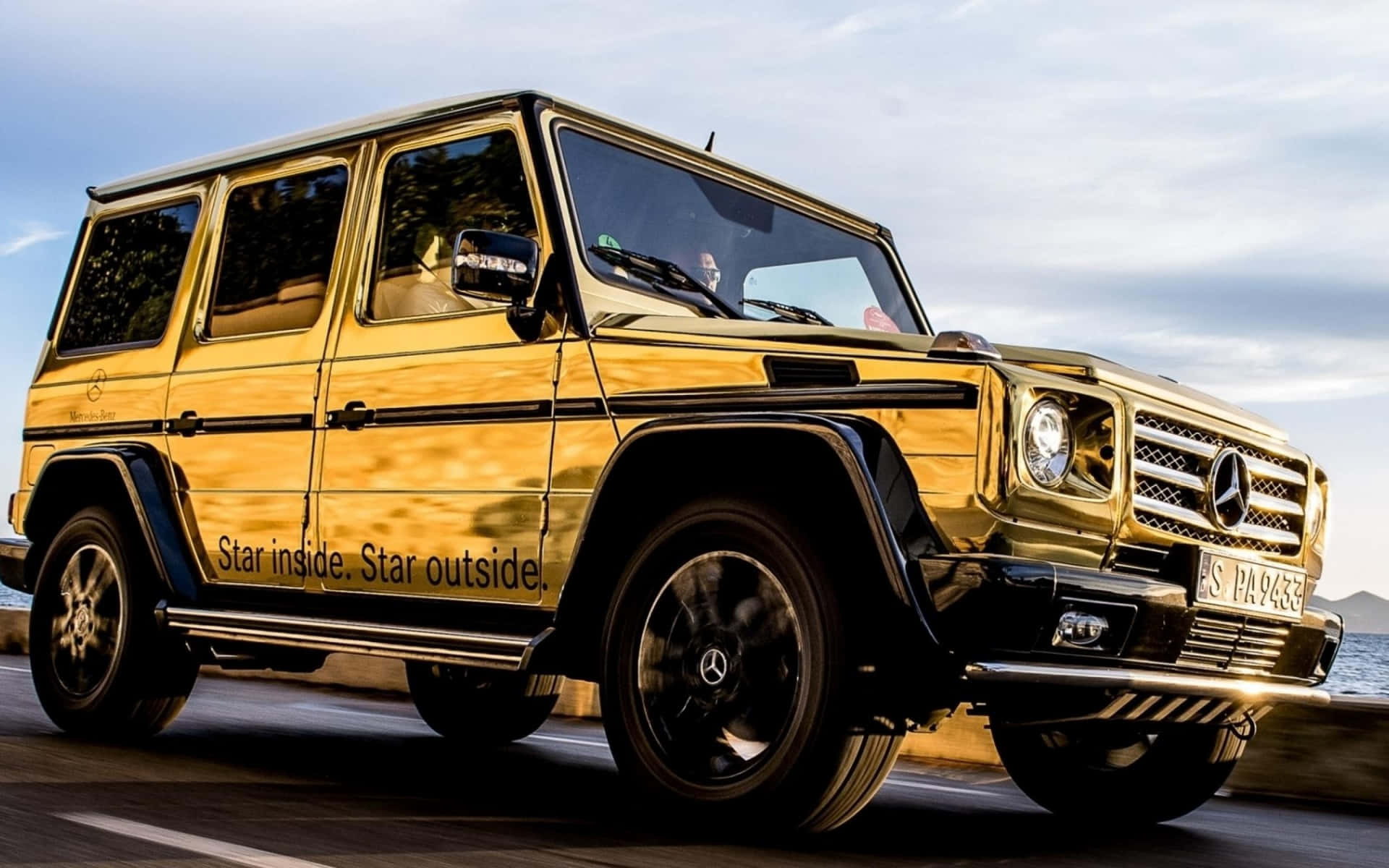 金色 Mercedes G-Class SUV 在海岸公路上行駛，背景是海洋。