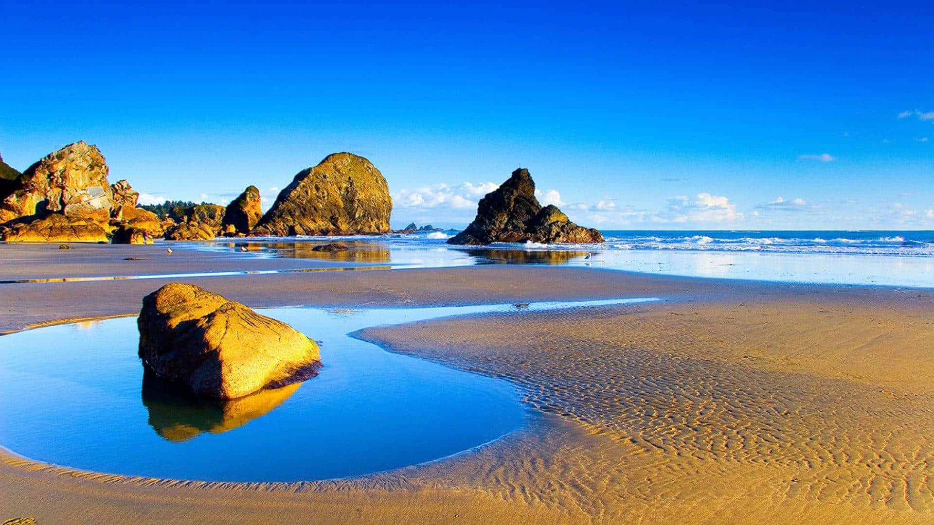 Seashore 桌布 壁紙 — 金色沙灘與碧綠海水相遇，環繞著巨大的海蝕柱，天空清澈湛藍，點綴著幾朵白雲。