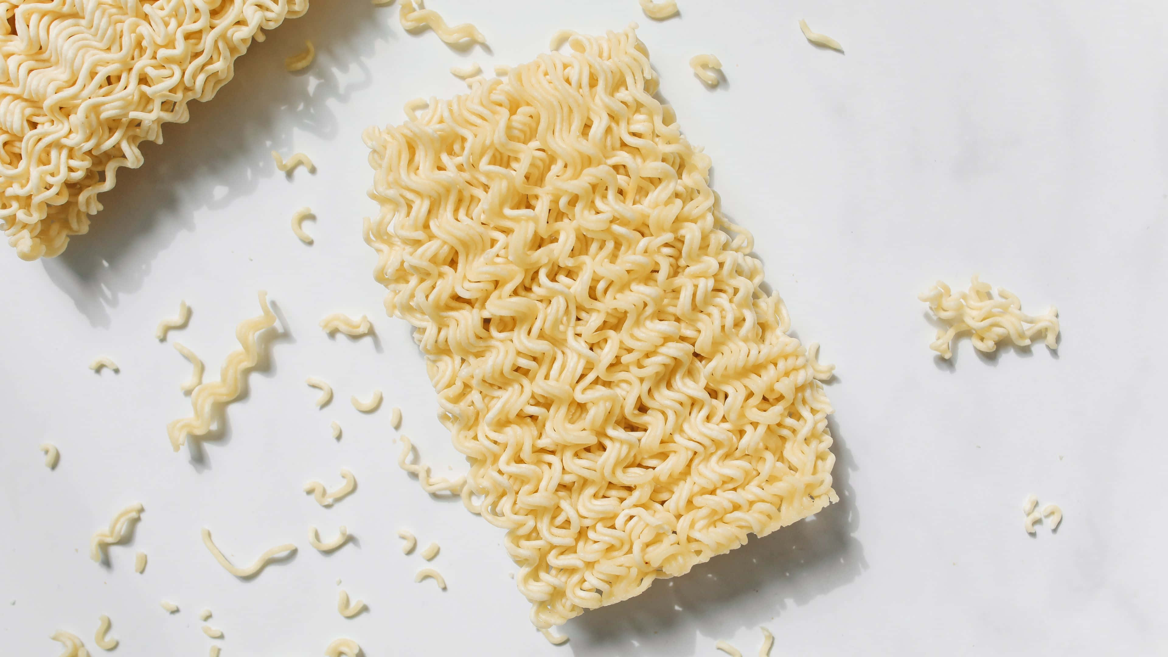 Noodles 桌布 壁紙 — 金色波浪形即食拉麵的特寫，散落的麵條在乾淨的白色大理石表面上。