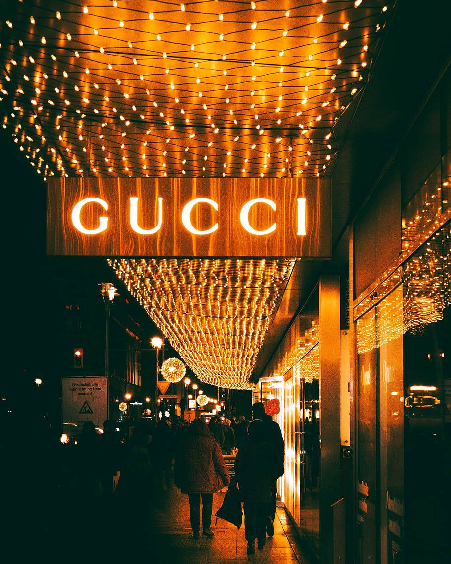 Gucci 桌布 壁紙 — 金色燈串拱門橫跨繁華的夜晚街道，通往發光的Gucci商店招牌。