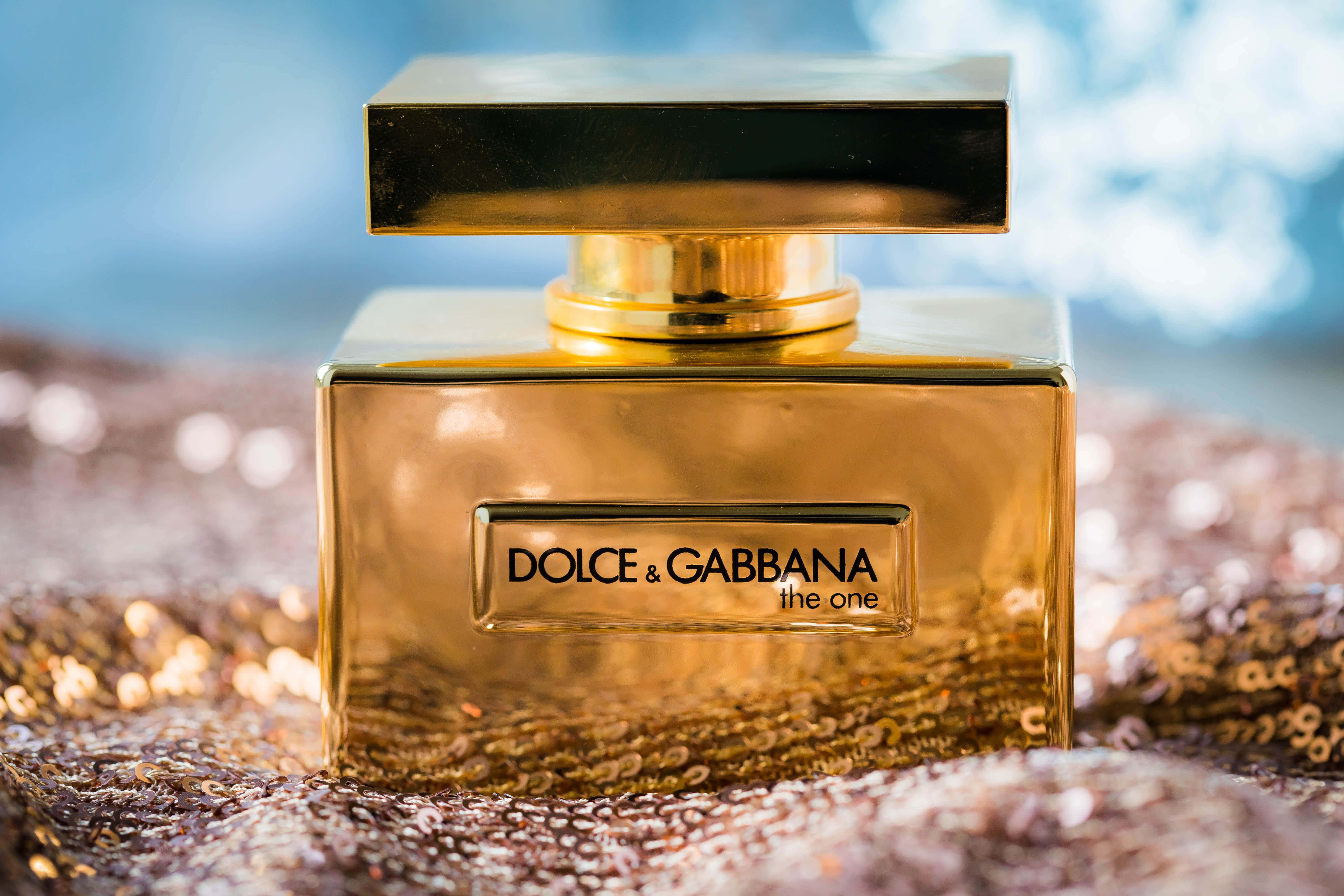 Fragrance 桌布 壁紙 — 金色的 Dolce & Gabbana 香水瓶在閃閃發光的亮片表面上閃耀，背景是柔和的藍色散景。