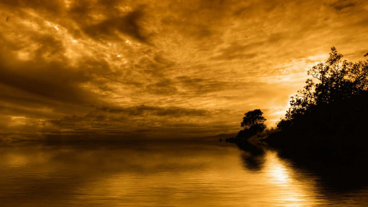 1280x720 桌布 壁紙 — 金色的夕陽在戲劇性的雲層下，將溫暖的光線投射在平靜的湖面和沿岸的樹木剪影上。