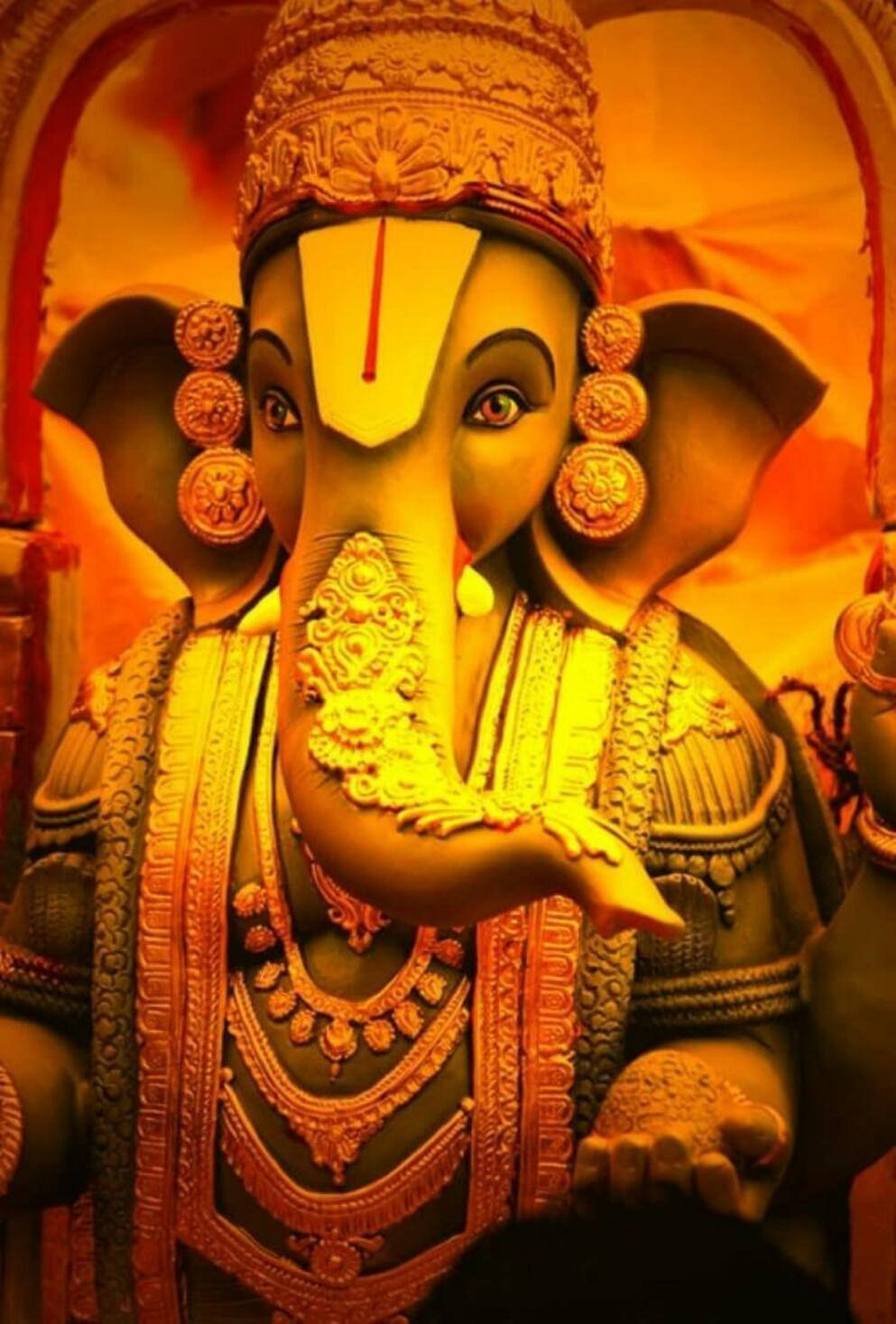ganesh iPhone 桌布 壁紙 — 金色的象神雕像，飾以精緻珠寶和皇冠，在耀眼的橙色背景下溫暖發光。