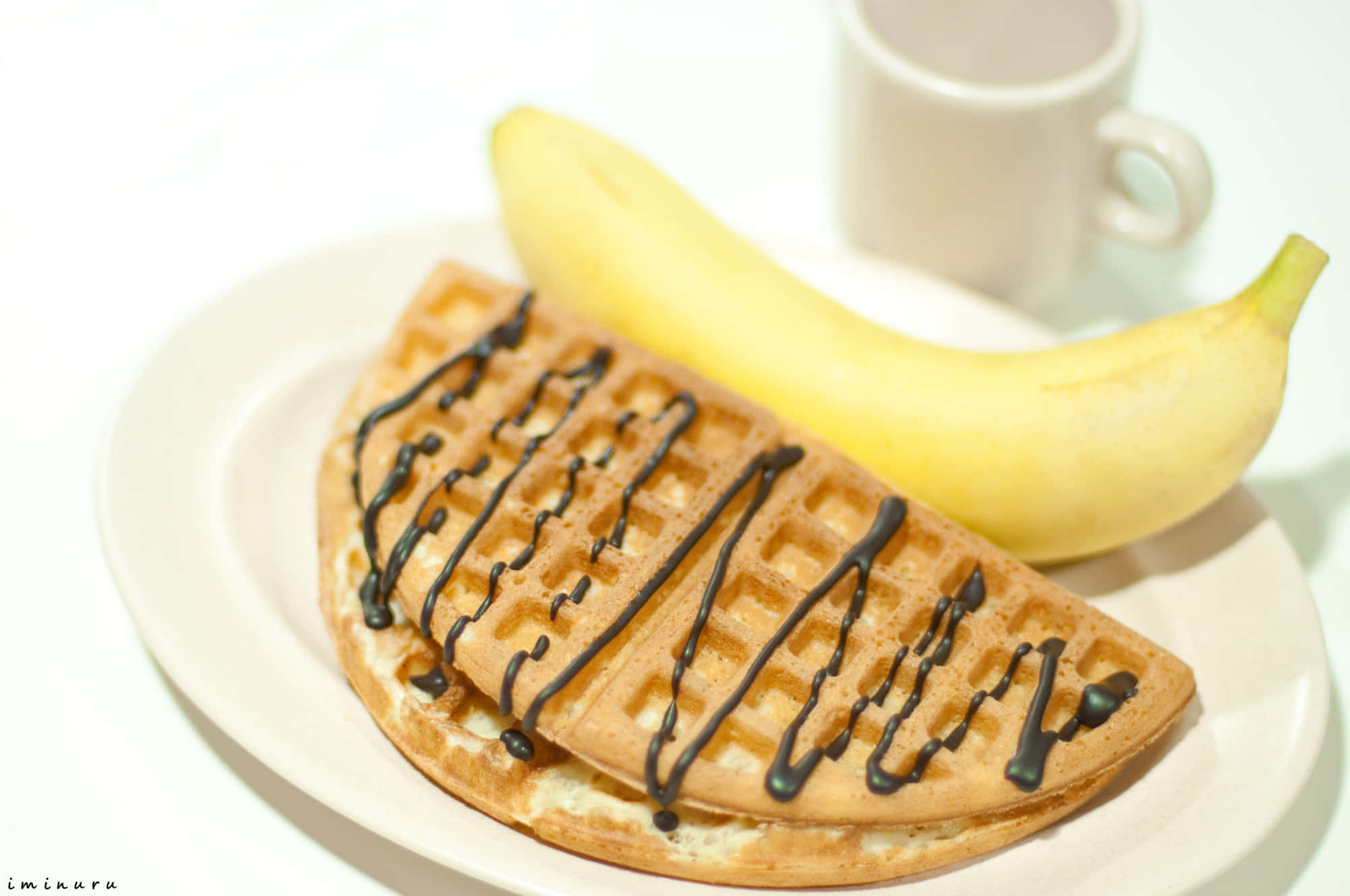 Waffle 桌布 壁紙 — 金色窩夫淋上朱古力醬，旁邊放著一條熟香蕉，放在白色碟上，背景有杯模糊的杯子。
