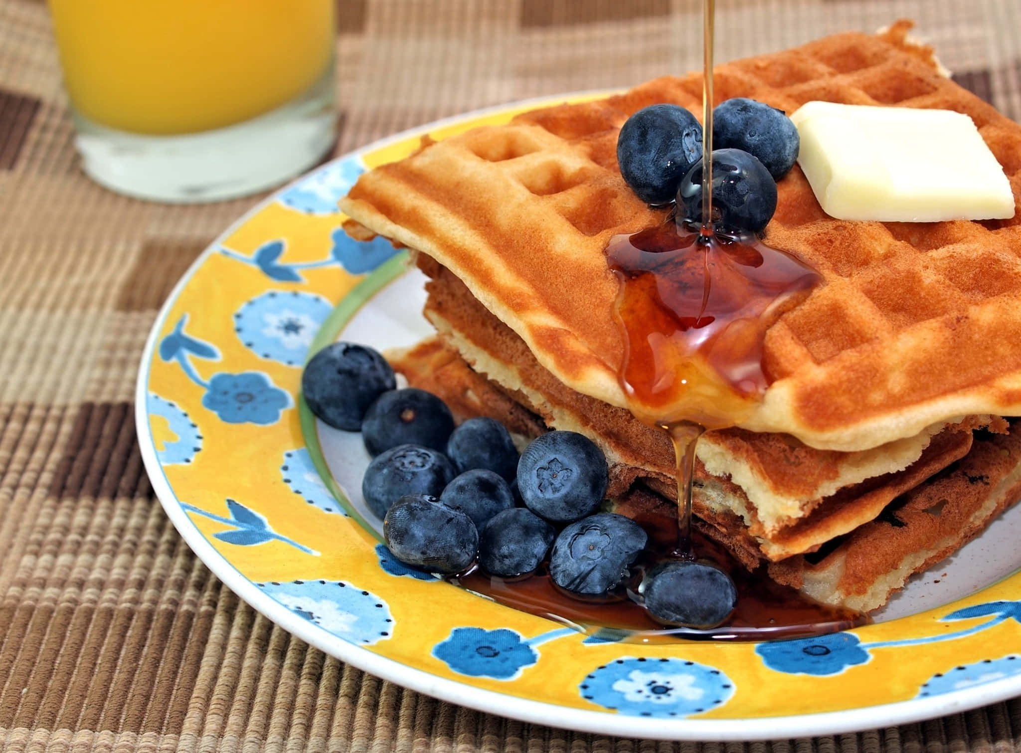 Waffle 桌布 壁紙 — 金色窩夫高高疊起，配上糖漿、藍莓和牛油，放在花卉圖案碟上，旁邊有橙汁。