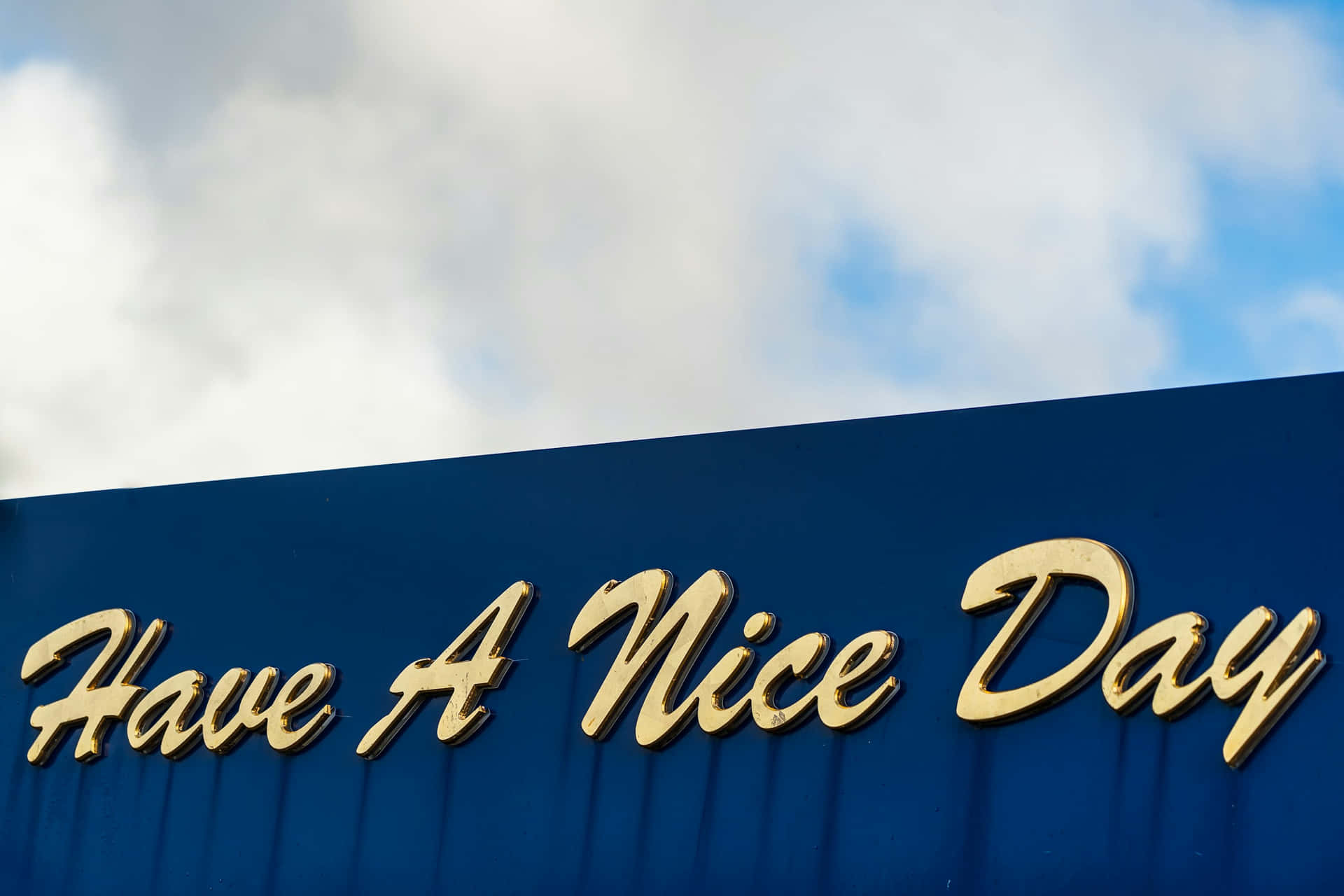  壁紙 — 金色草書字母在深藍色標誌上拼出「Have A Nice Day」，背景是局部多雲的天空。