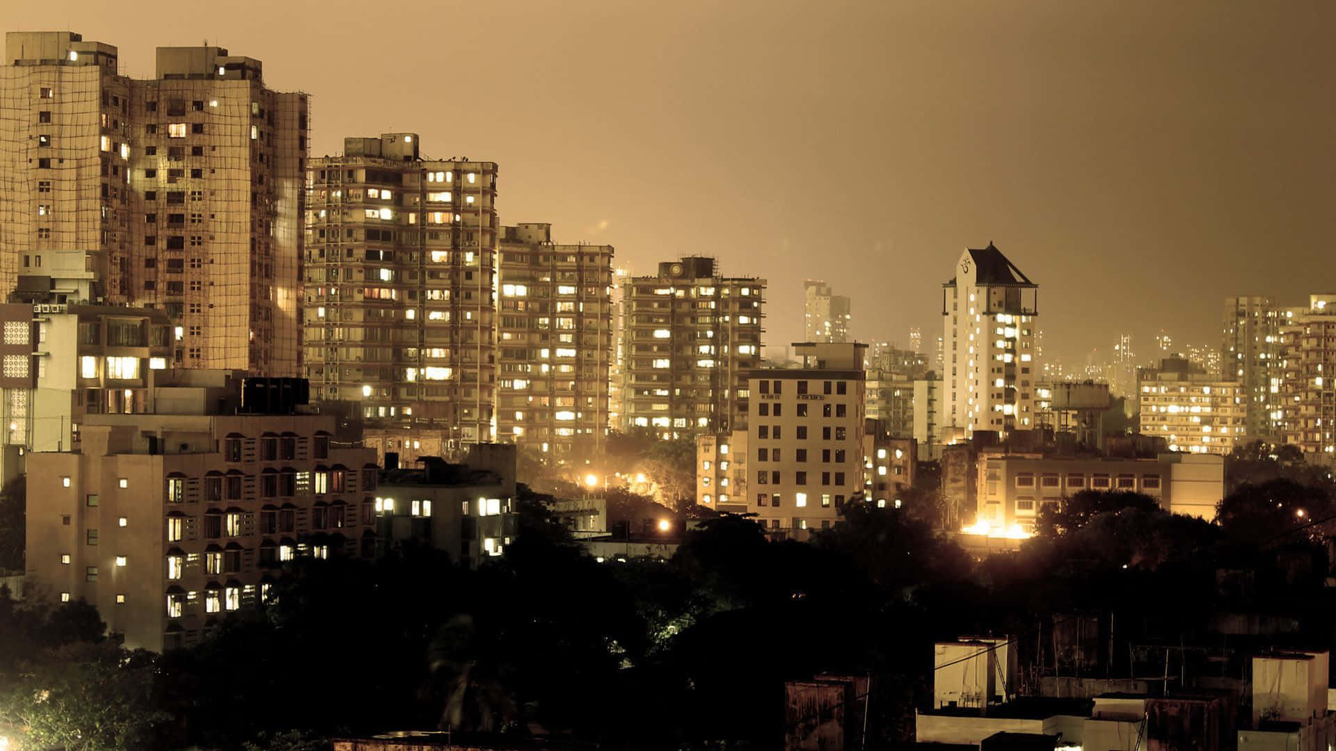 Mumbai 桌布 壁紙 — 金色調的夜景城市景觀，高樓大廈燈火通明，前景有深色樹木剪影。