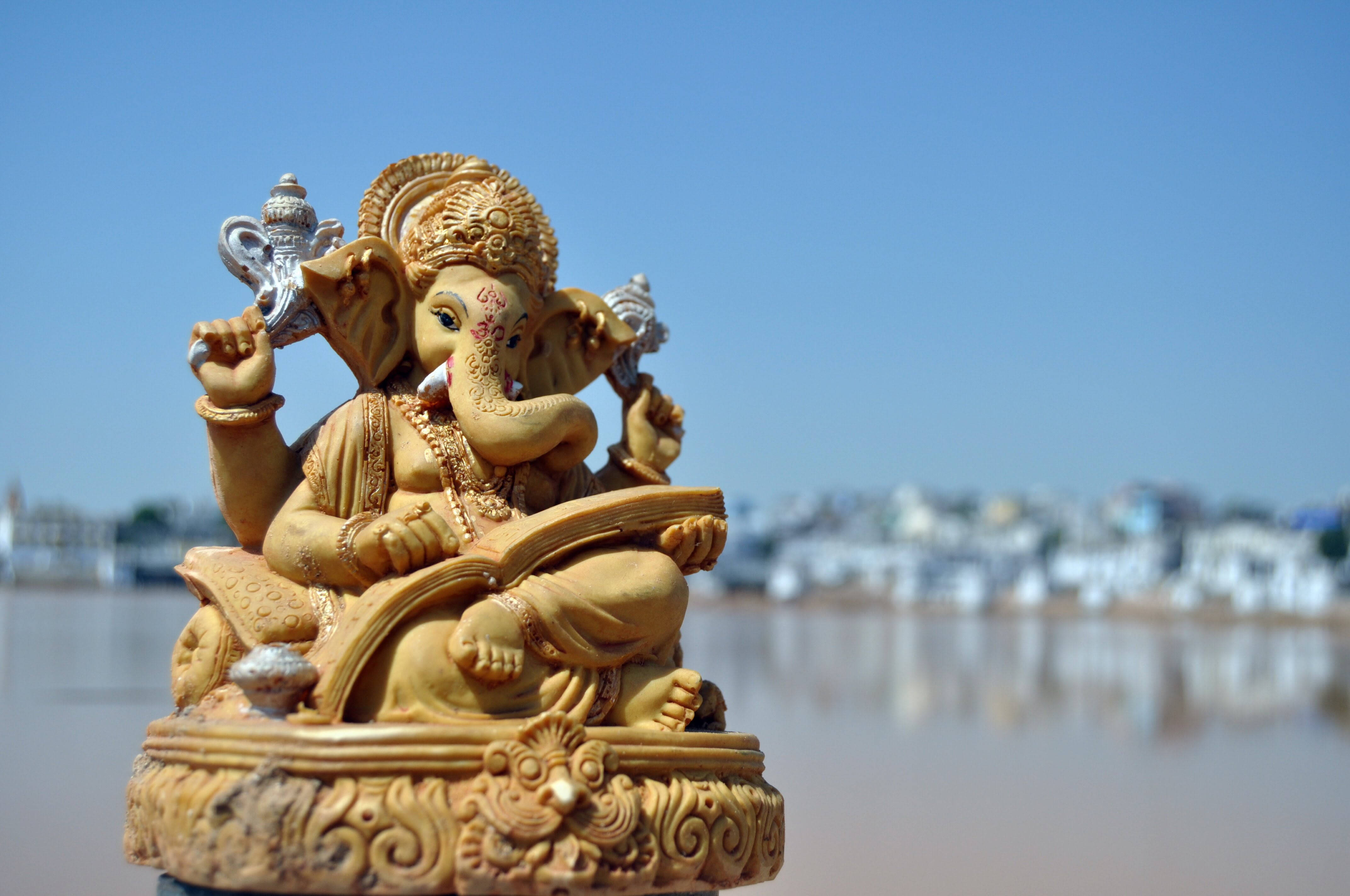 Hindu God 桌布 壁紙 — 金色象神雕像靜靜地坐在河邊，模糊的城市景觀在清澈的藍天下。