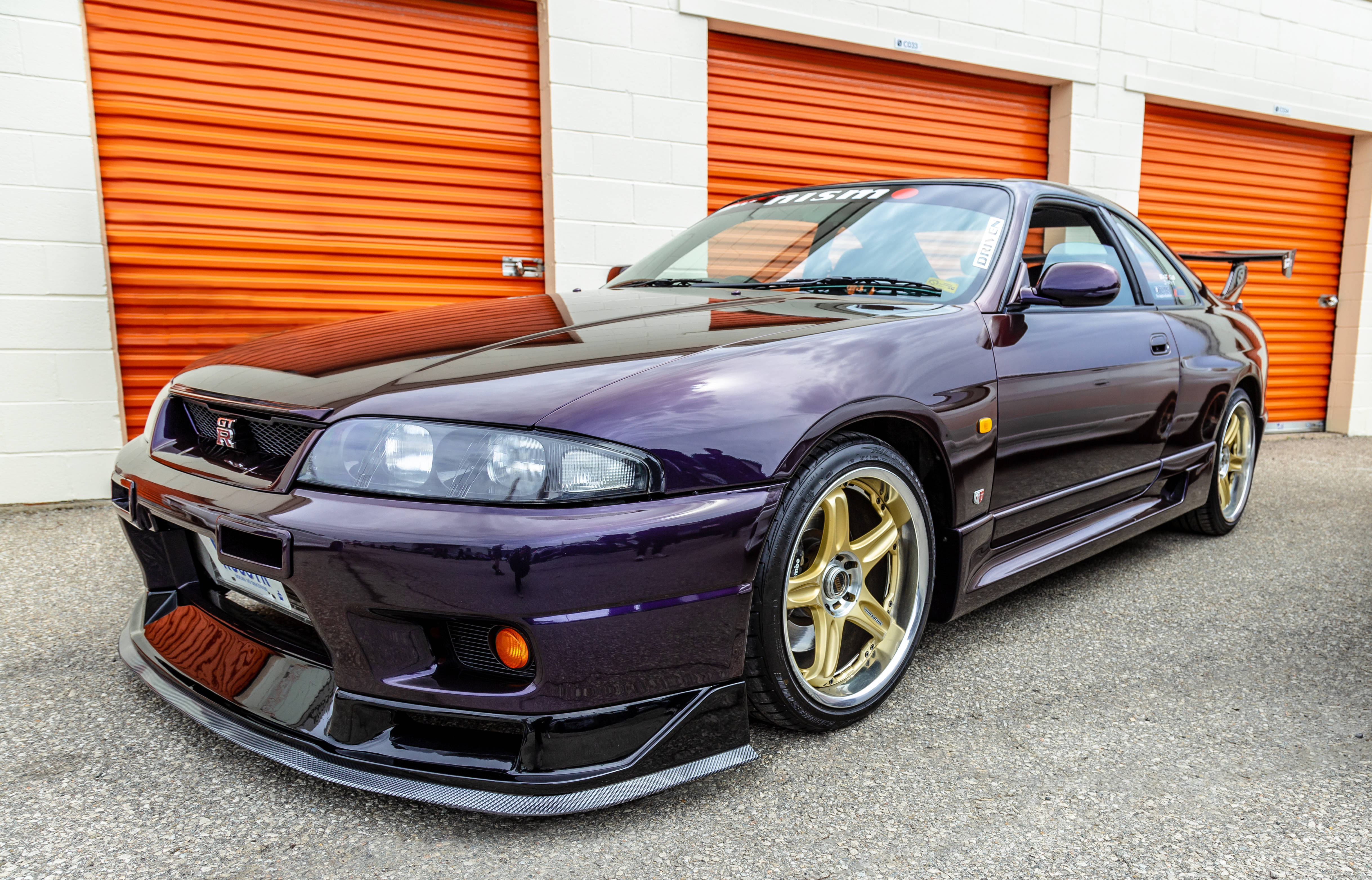 金色輪圈尼桑GTR 壁紙 — 一輛光澤紫色日產Skyline R34跑車配金色輪圈，停在亮橙色車庫門前。