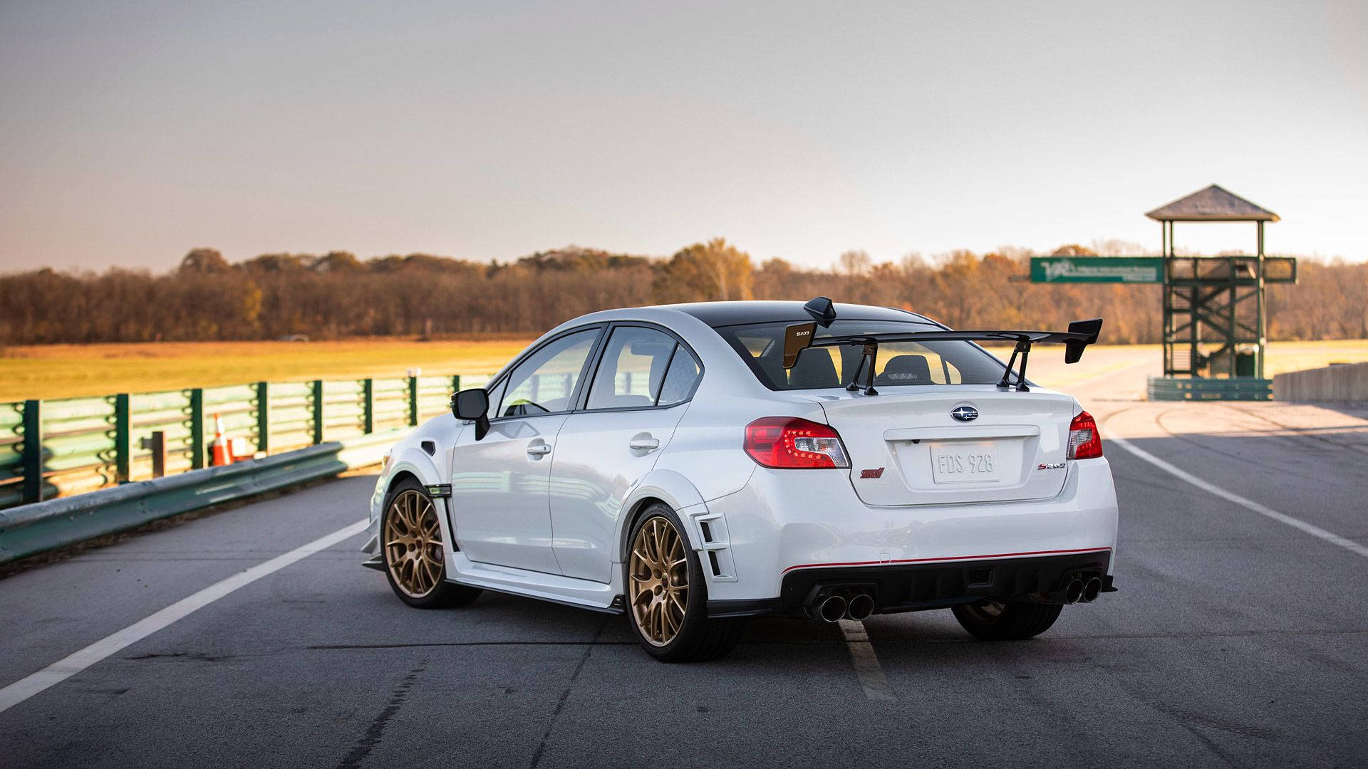 金色輪圈斯巴魯 壁紙 — 一輛白色Subaru WRX STI配備金色輪圈及大型尾翼，在黃金時段的賽道上蓄勢待發。