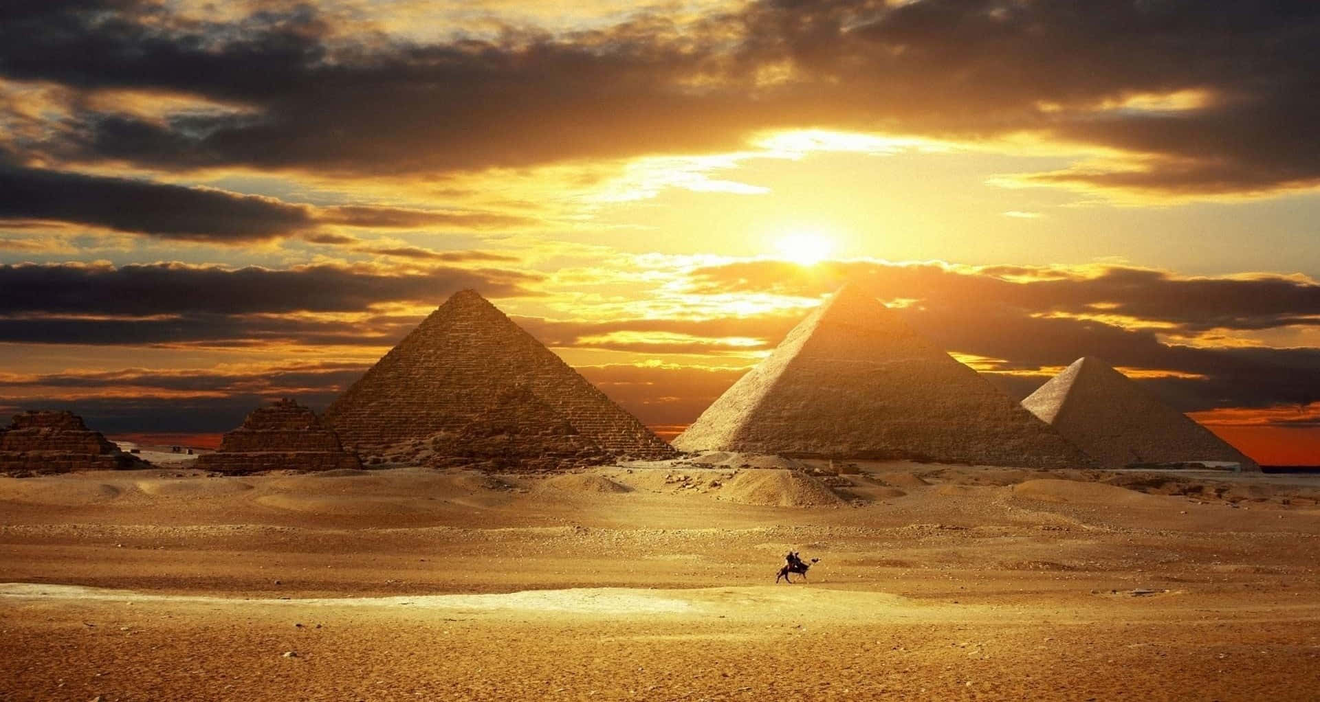 Egypt 桌布 壁紙 — 金色金字塔從沙漠沙地中升起，一名孤獨的騎手騎著駱駝在戲劇性的日落天空下穿越沙丘。