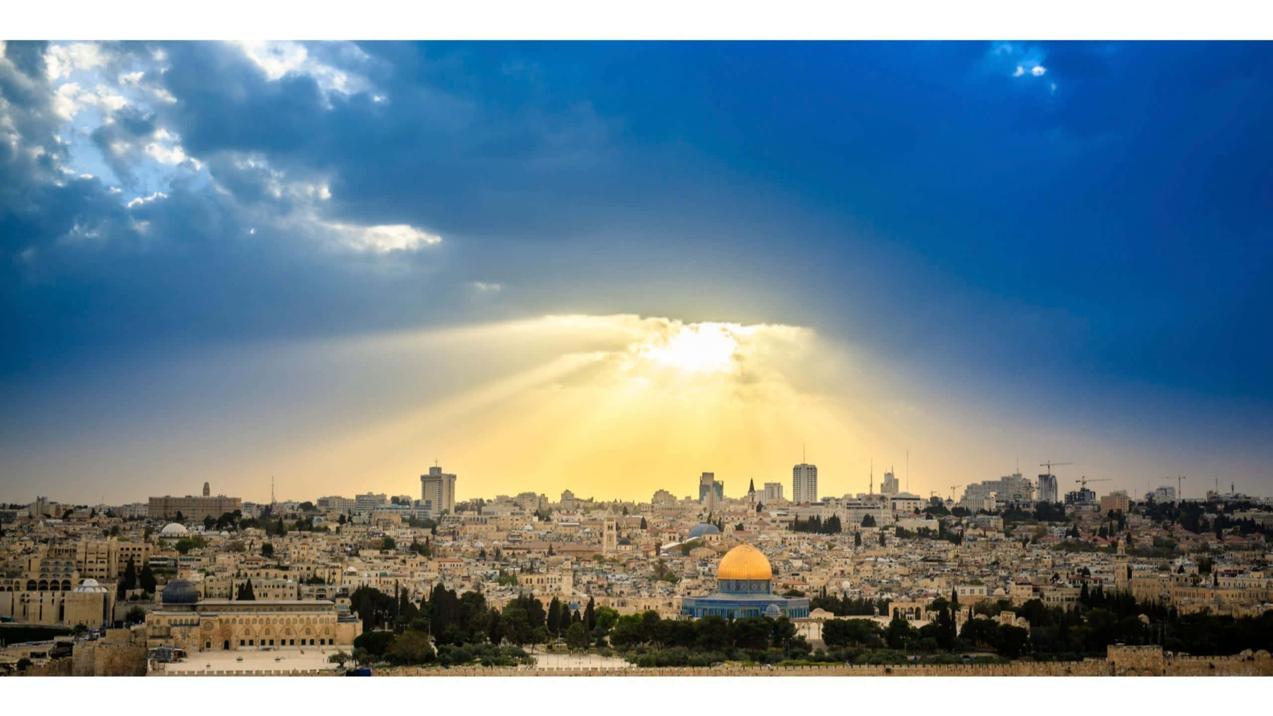 Israel 桌布 壁紙 — 金色陽光穿過戲劇性的雲層，照耀在耶路撒冷古老的石頭城市景觀上，圓頂清真寺在溫暖的光線中閃耀。