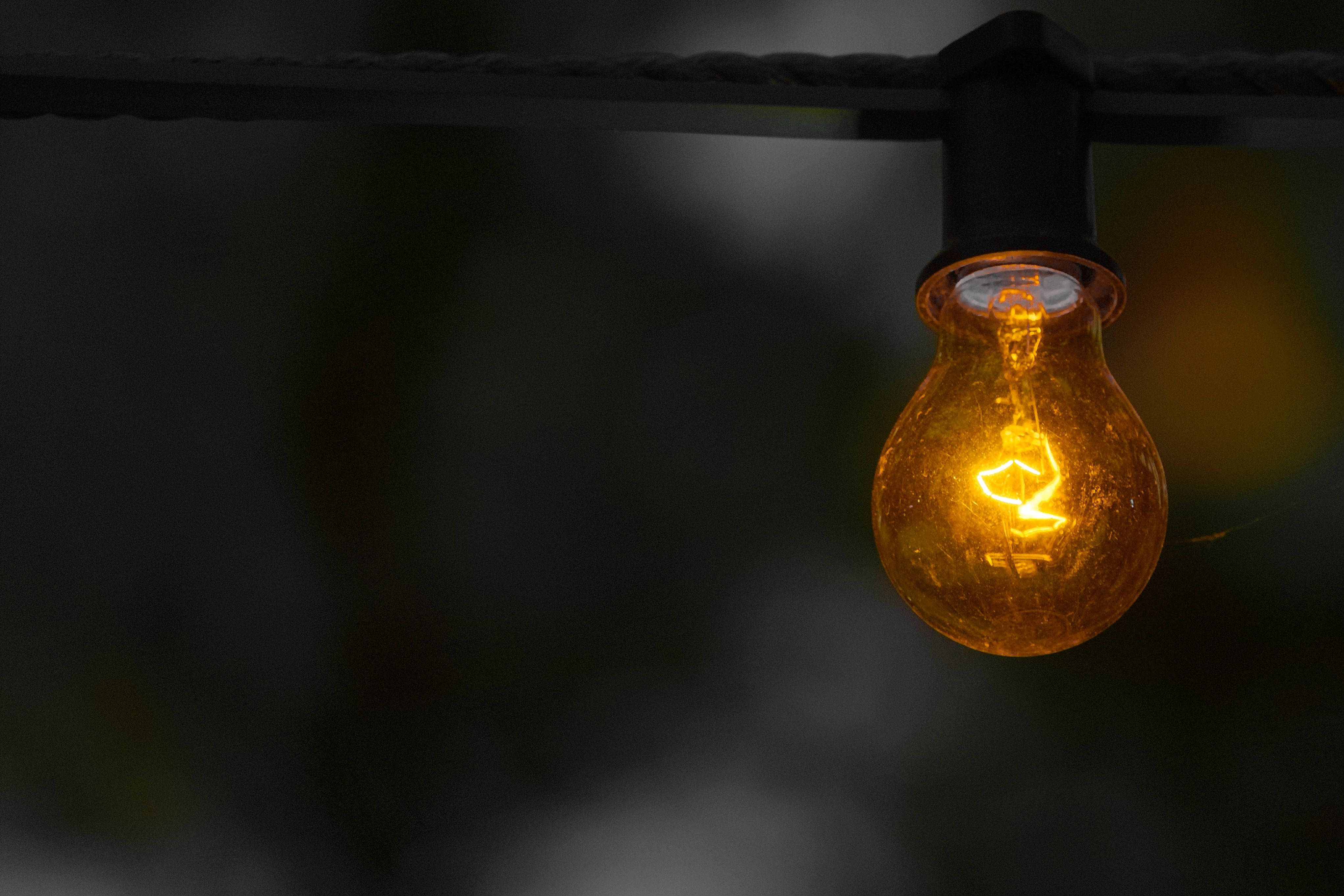 金色電力燈泡 壁紙 — 一個發光的琥珀色燈絲燈泡掛在黑色電線上，背景是黑暗模糊的。