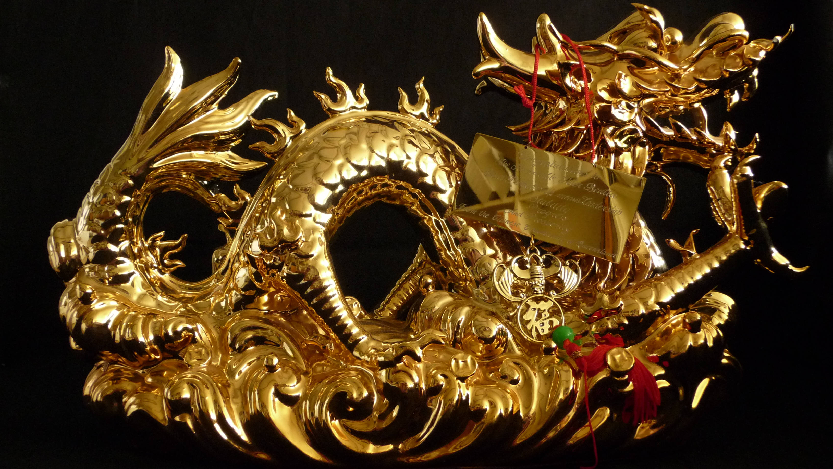 金色龍擺件 壁紙 — 閃閃發光的金色龍雕塑在黑色背景上動態盤繞，飾有紅色流蘇和幸運符。