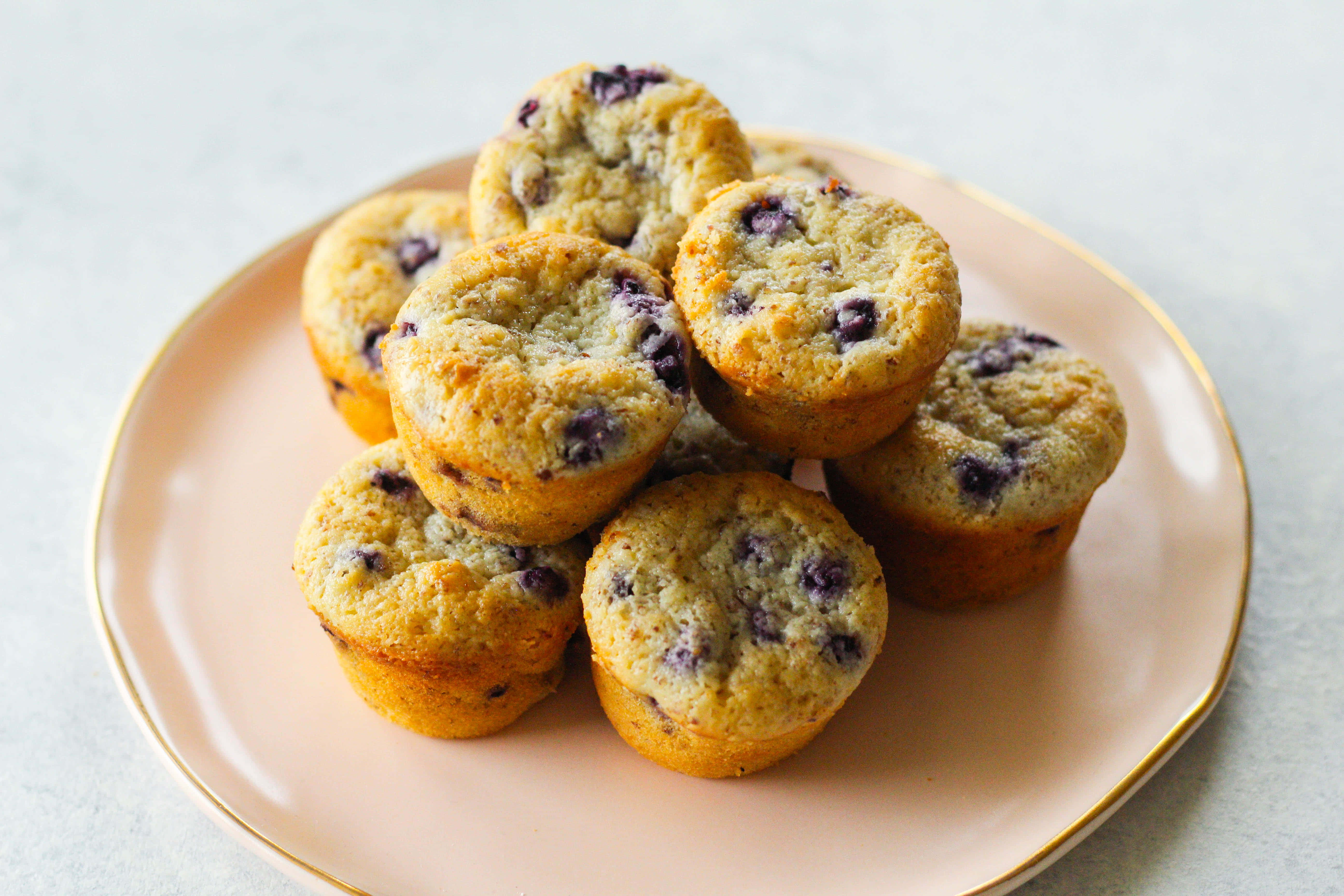 Blueberry Muffins 桌布 壁紙 — 金藍莓鬆餅高高堆放在柔軟的粉紅色金邊碟子上，背景為淺灰色。