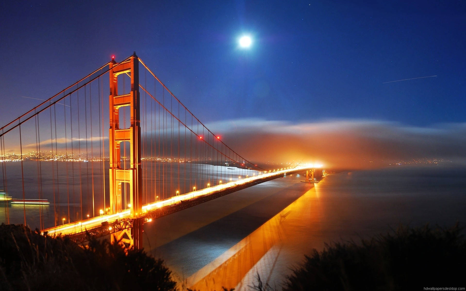 金門大橋夜景最佳桌面壁紙 壁紙 — 金門大橋在明亮的月亮下溫暖地發光，映襯著深藍色的夜空，城市燈光在下方閃爍。