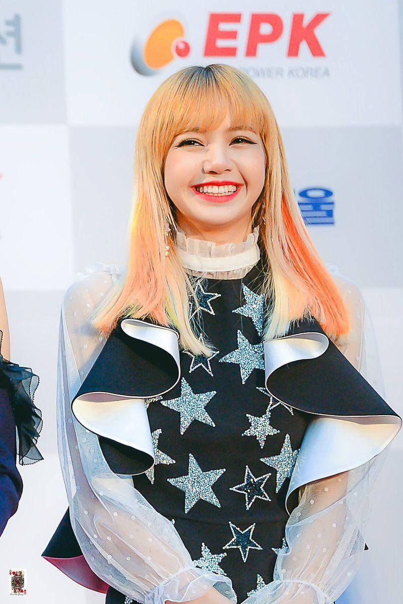 金髮BLACKPINK LISA 笑容燦爛 壁紙 — 一位金髮紅唇、面帶微笑的女性，在記者會上穿著帶有星星圖案的黑色連衣裙和透明荷葉邊袖。