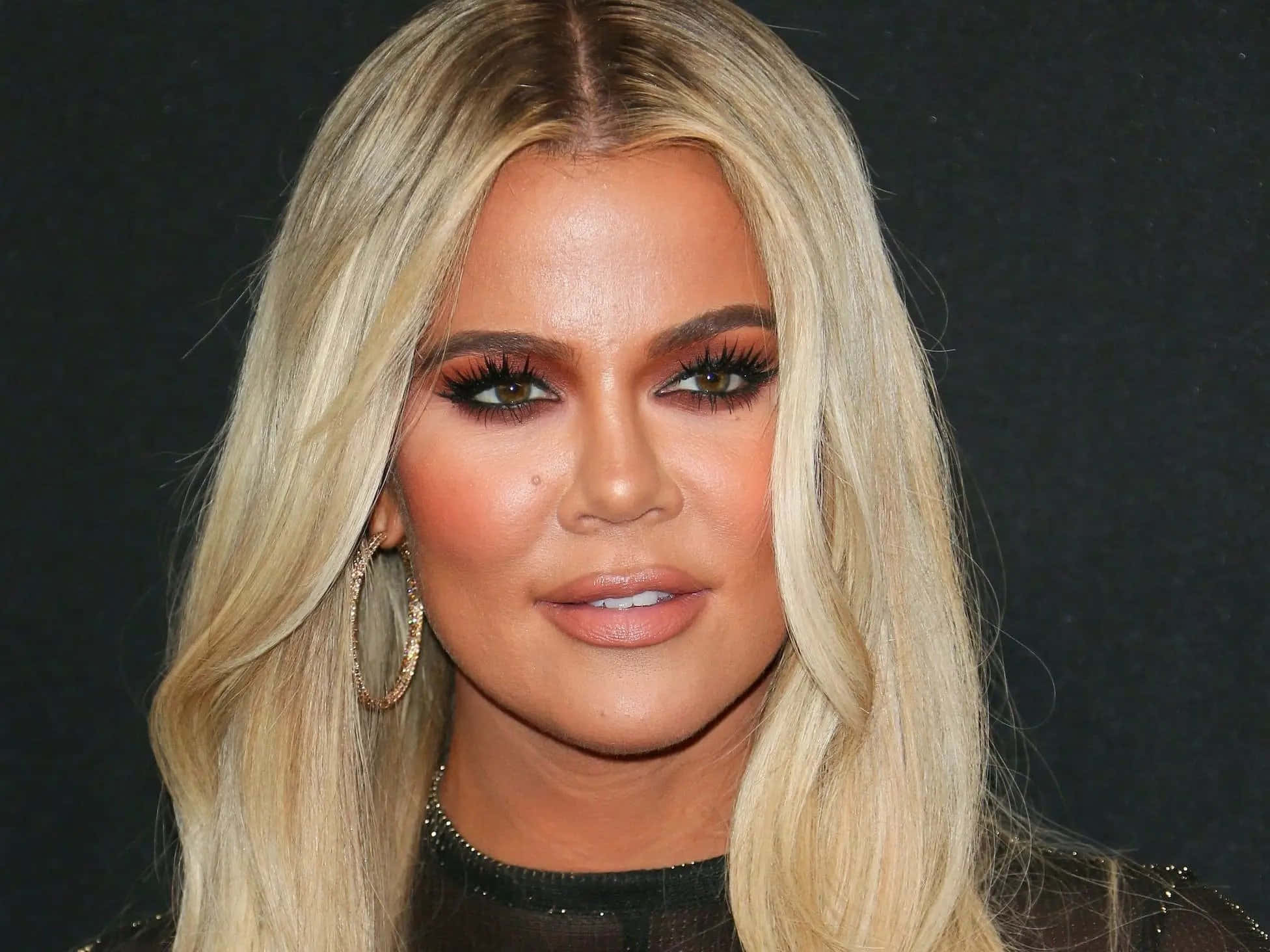 Khloe Kardashian 桌布 壁紙 — 金髮女子特寫肖像，妝容大膽，戴著金色圓環耳環，背景深色。