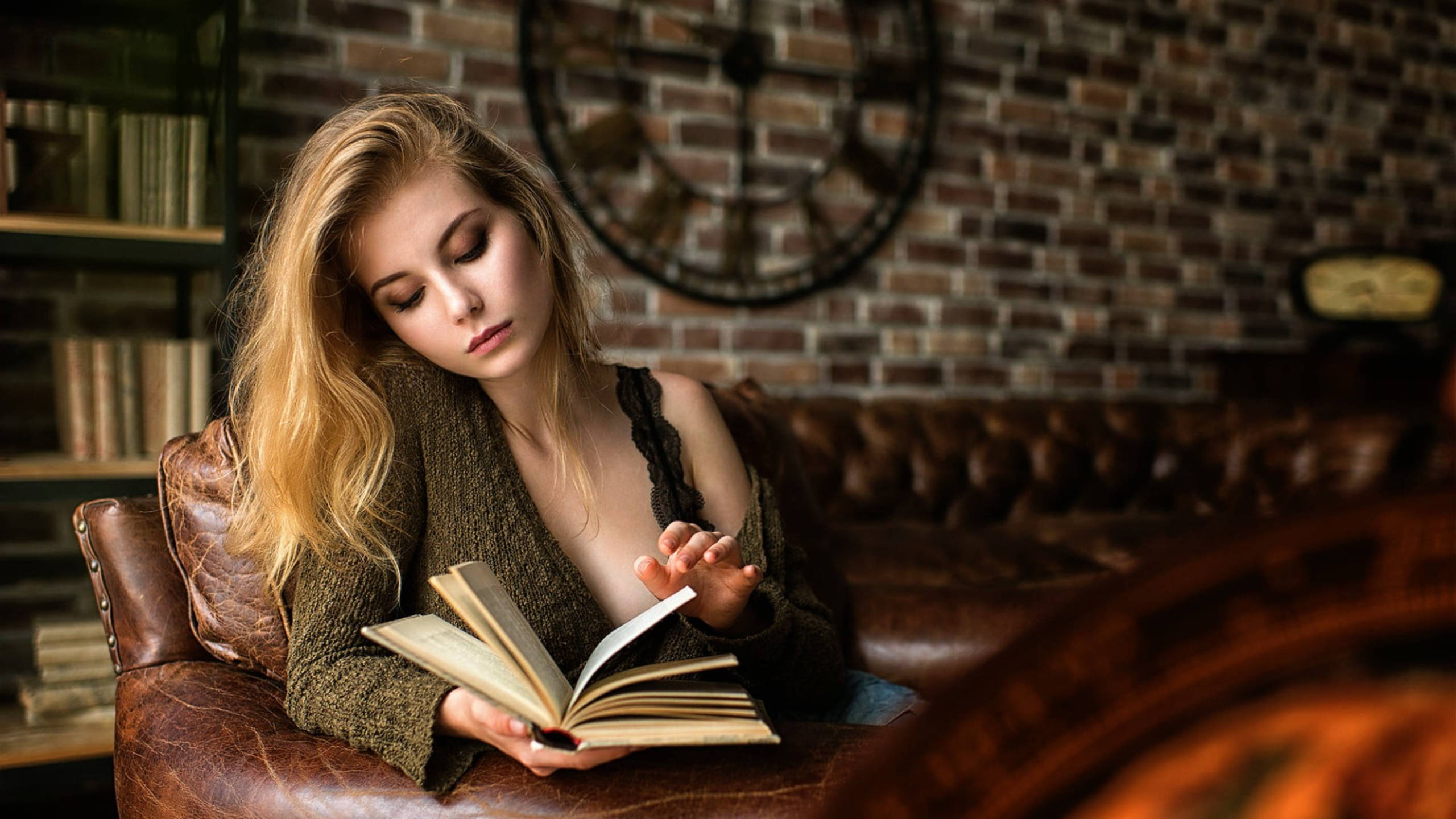 金髮女孩閱讀書籍 壁紙 — 一位金髮年輕女性坐在皮革扶手椅上，在一個有磚牆、書架和大時鐘的舒適房間裡看書。
