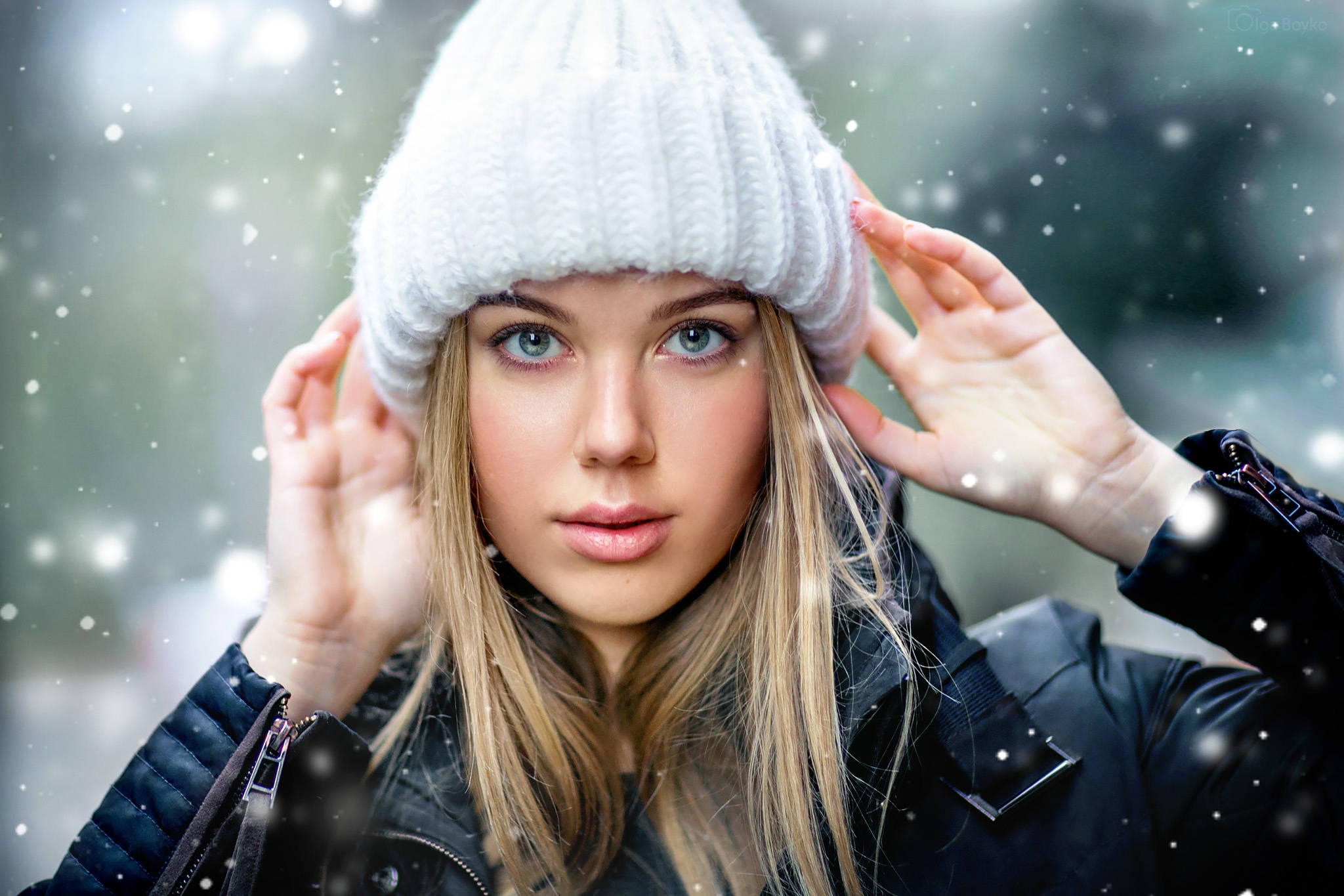 金髮女模特戴毛線帽雪景 壁紙 — 一位年輕女性，擁有引人注目的藍色眼睛和金色頭髮，戴著白色針織冷帽和黑色外套，雪花輕輕飄落在她周圍的冬日場景中。