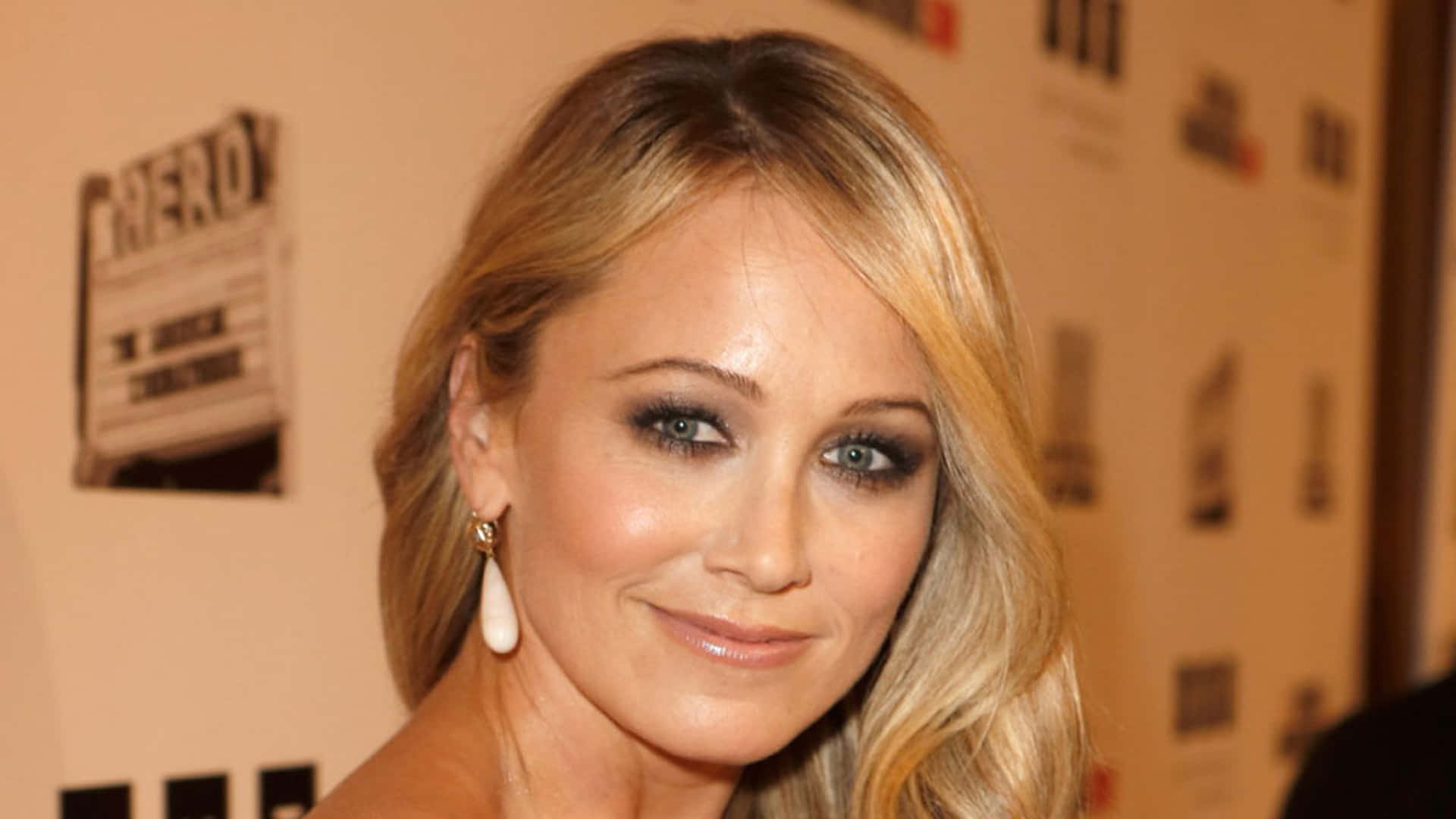 Christine Taylor 桌布 壁紙 — 金髮碧眼、戴著珍珠耳環的女性在品牌背景前對著鏡頭微笑的特寫肖像。