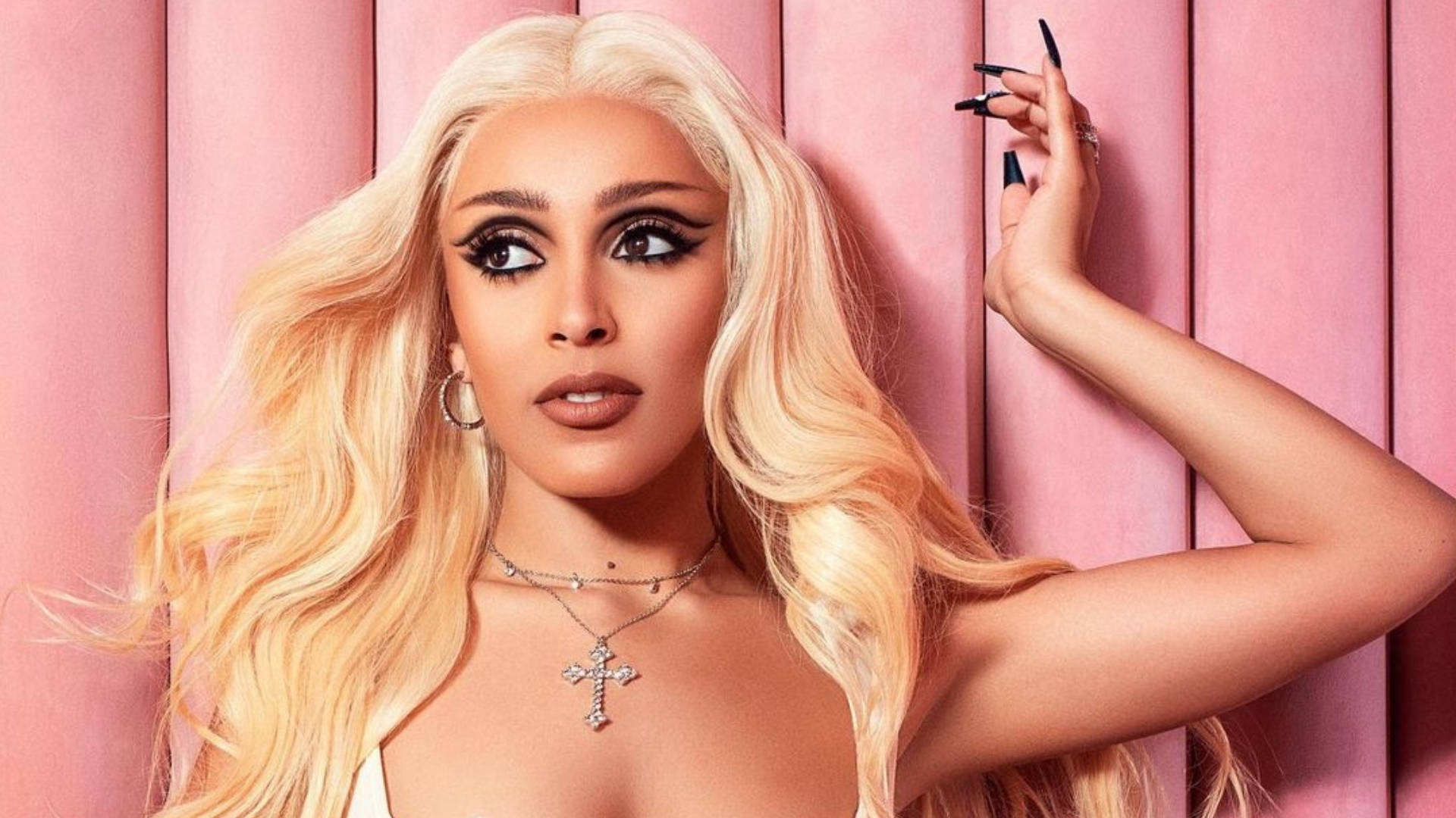 金髮魅力Doja Cat 壁紙 — 一位金髮長髮、妝容大膽的迷人女性，在柔和的粉紅色鑲板牆前擺姿勢。