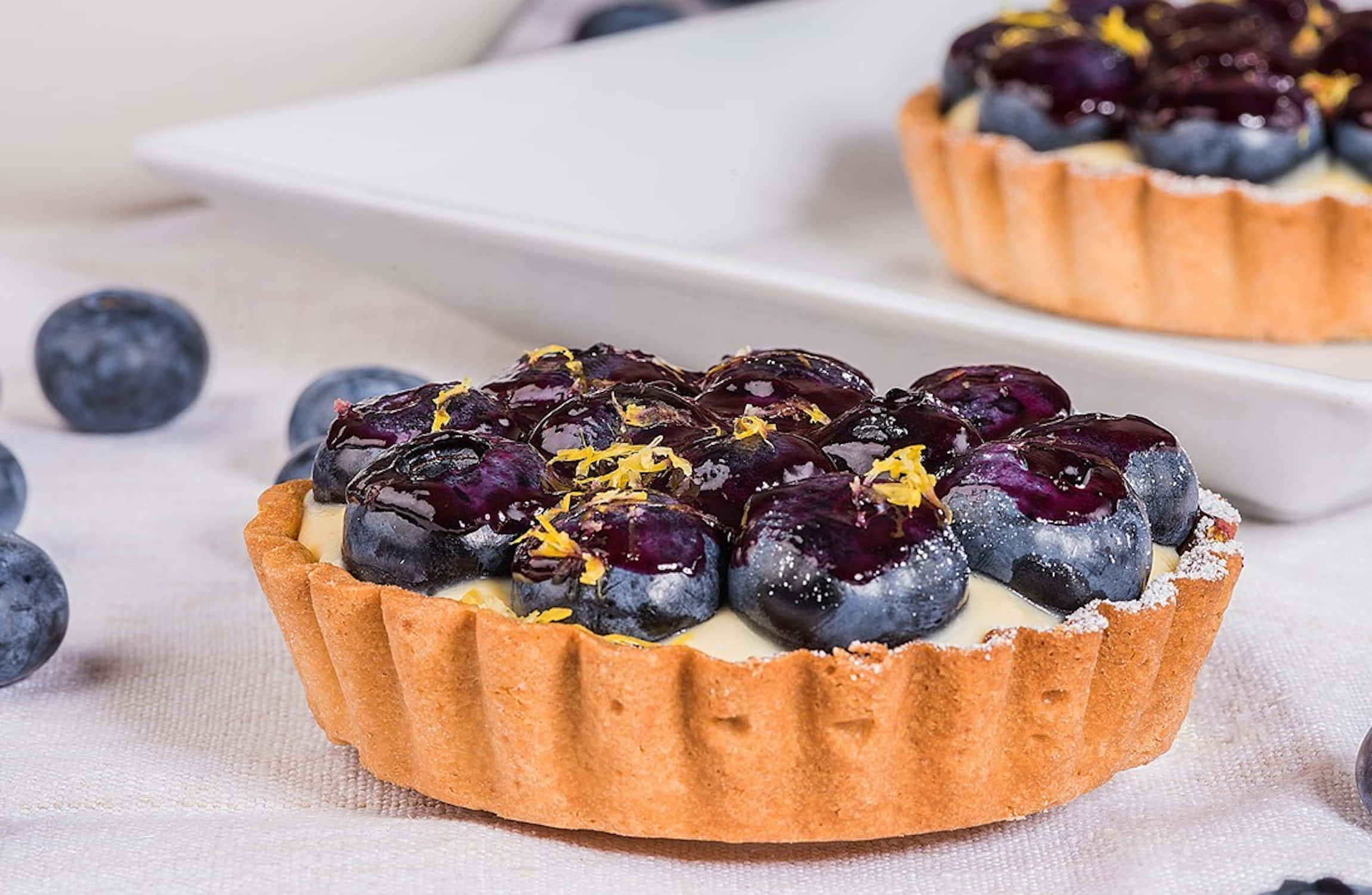 Blueberries Tart 桌布 壁紙 — 金黃色撻的近鏡，內餡充滿忌廉吉士，上面鋪滿閃亮藍莓和檸檬皮。
