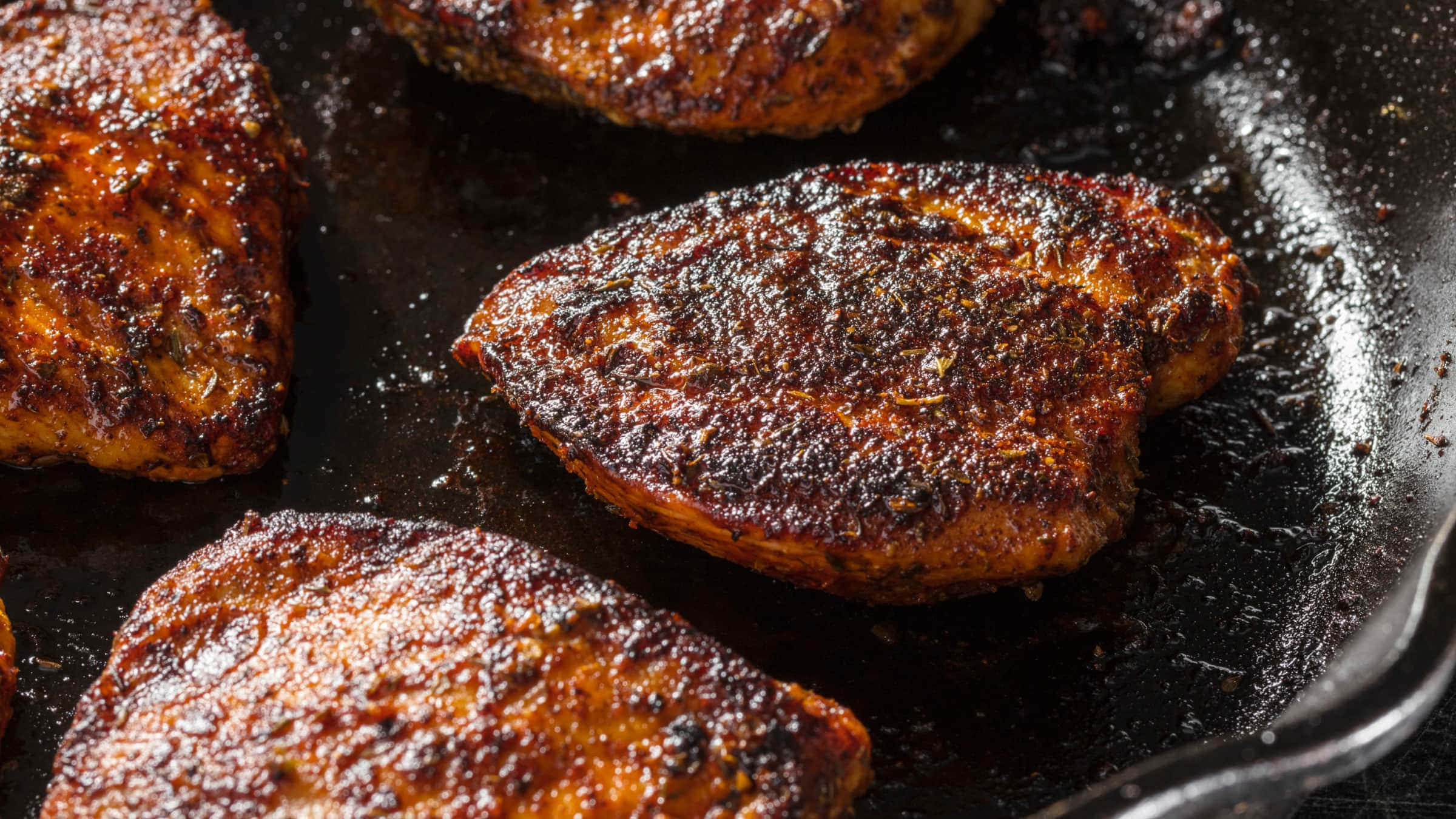 Blackened Chicken 桌布 壁紙 — 金黃色的烤雞腿在黑色的鑄鐵煎鍋中滋滋作響，可見調味料和油。