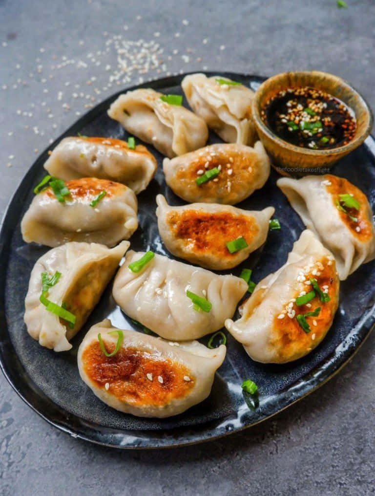 Local Food 桌布 壁紙 — 金黃色的煎餃，上面撒上蔥花和芝麻，放在深色碟子上，旁邊配上蘸醬。
