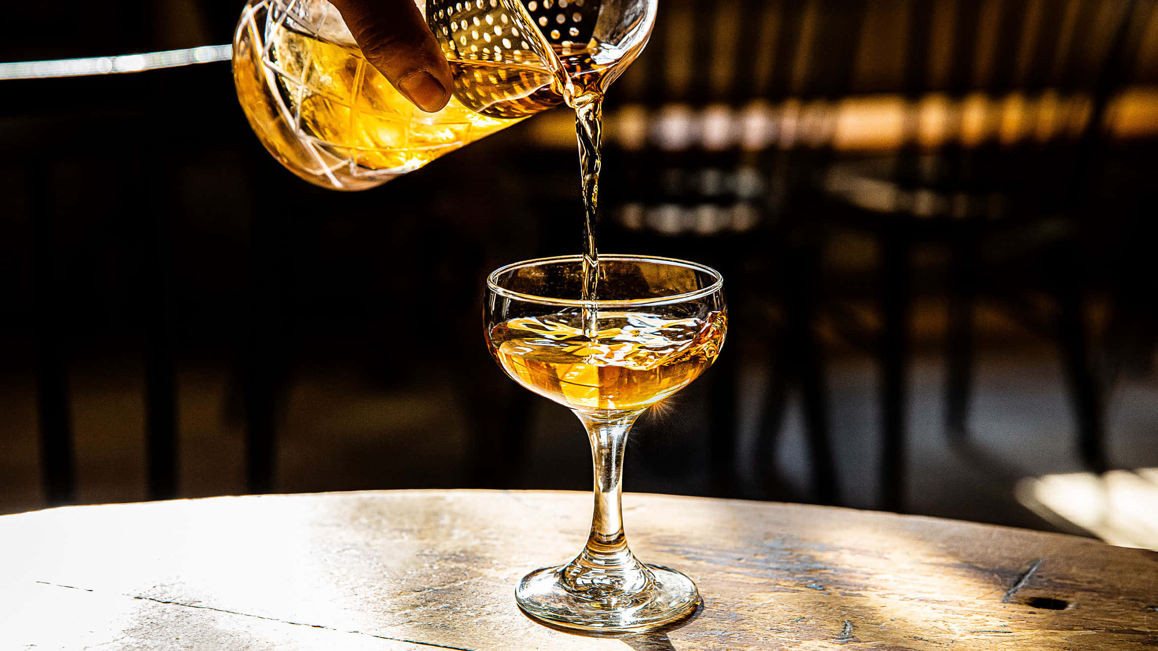 Beautiful Drink 桌布 壁紙 — 金黃色的琥珀色液體從玻璃雞尾酒調酒器倒入放在陽光照射的木桌上的碟形香檳杯。